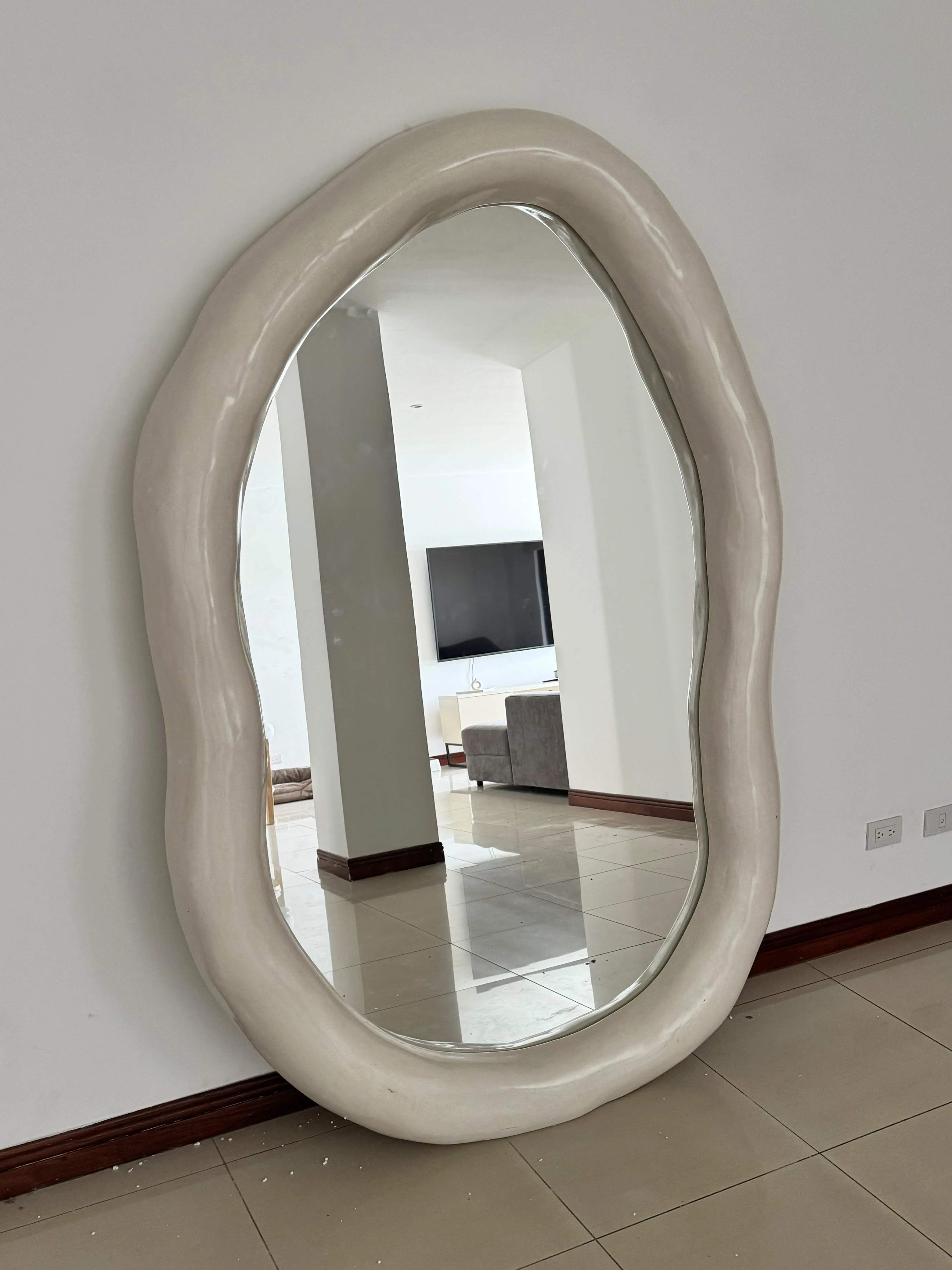 Espejo de forma orgánica ondulada con marco grueso de color crema, apoyado en una pared blanca sobre piso de cerámica