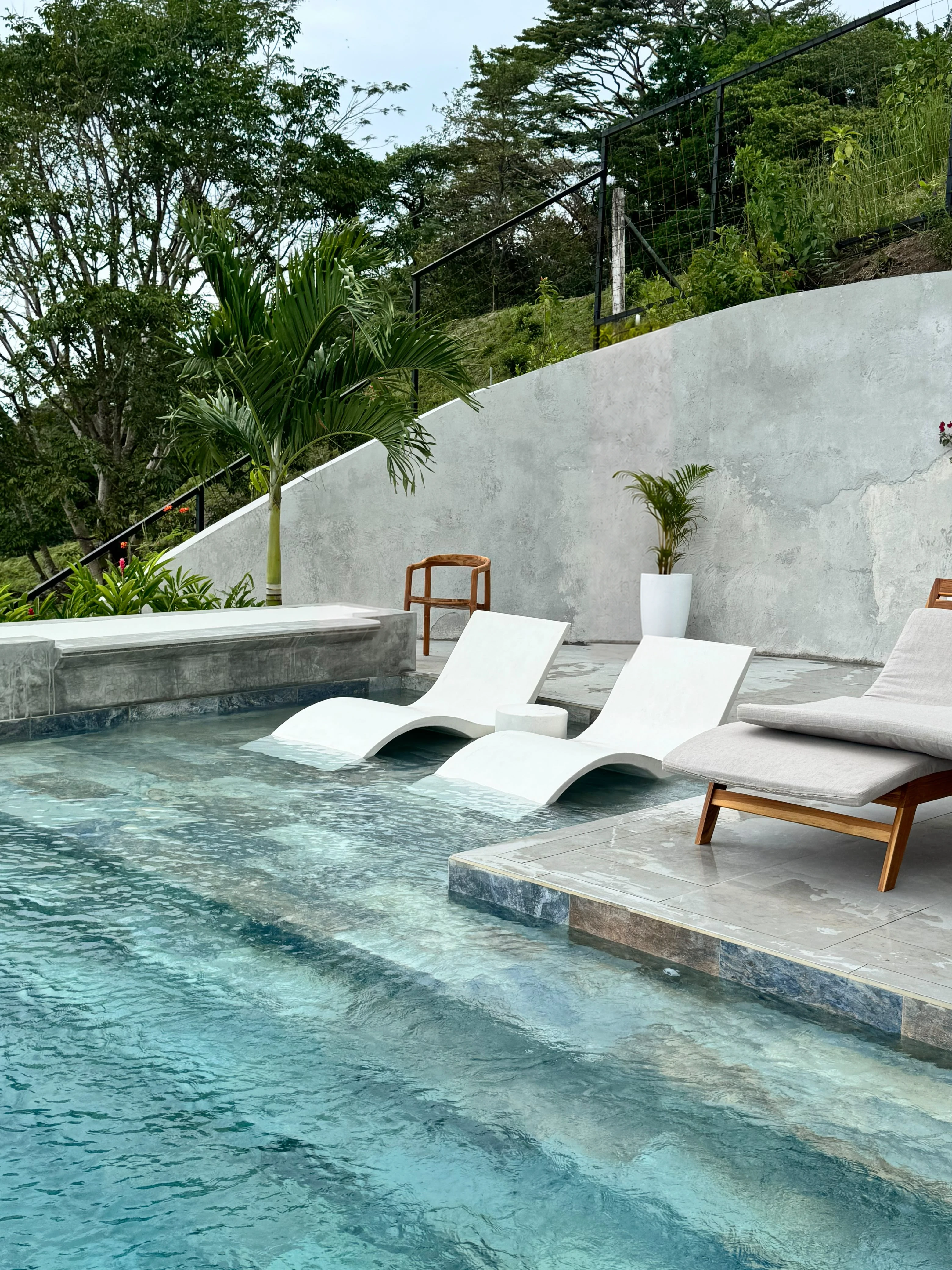 Tres camastros de concreto blanco con curvas onduladas dentro de una piscina, junto a una pared curva y plantas