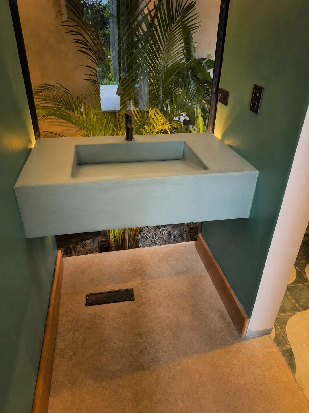 Lavatorio integrado flotante de concreto verde azulado con poza rectangular, grifería negra, en baño con paredes verdes y plantas