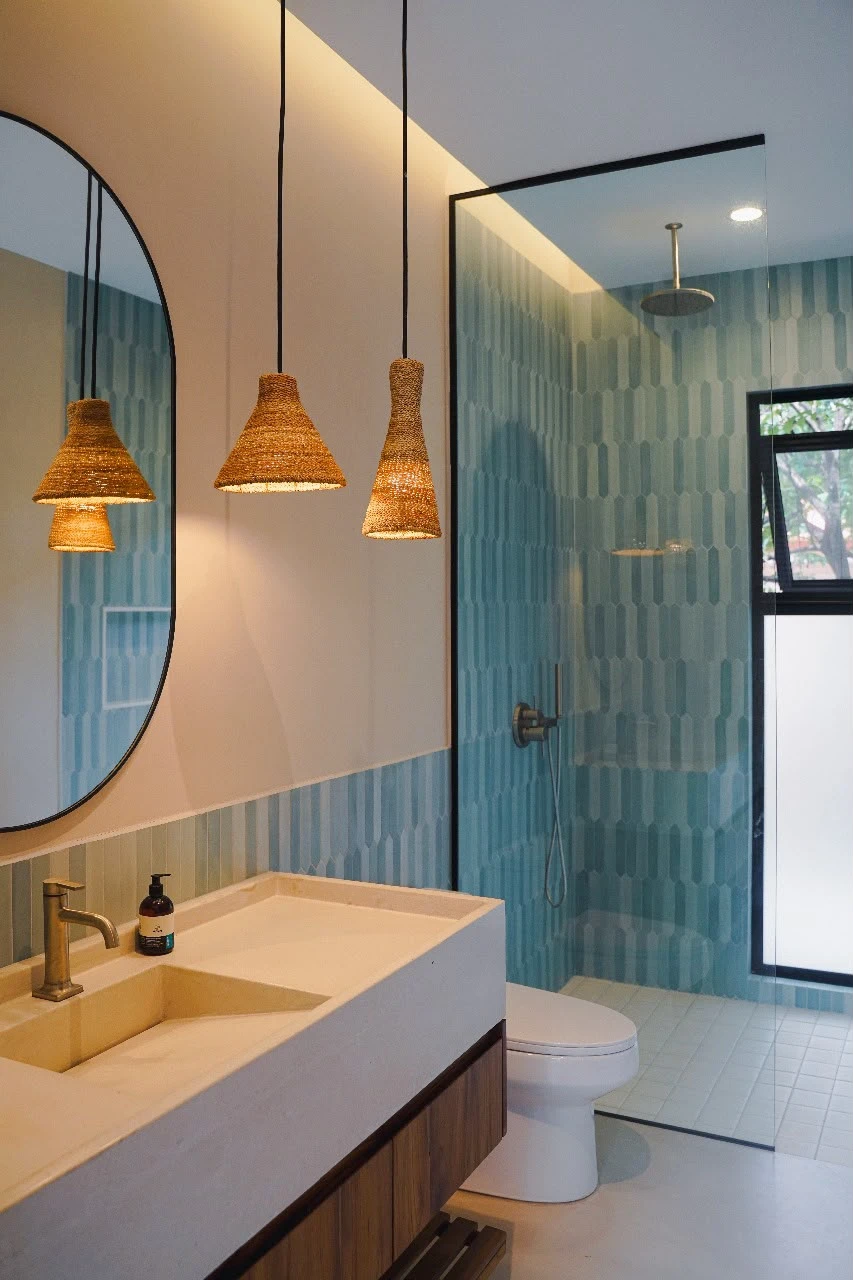 Lavatorio integrado de concreto claro con poza alargada y grifería dorada, junto a ducha con azulejos verdes y lámparas colgantes