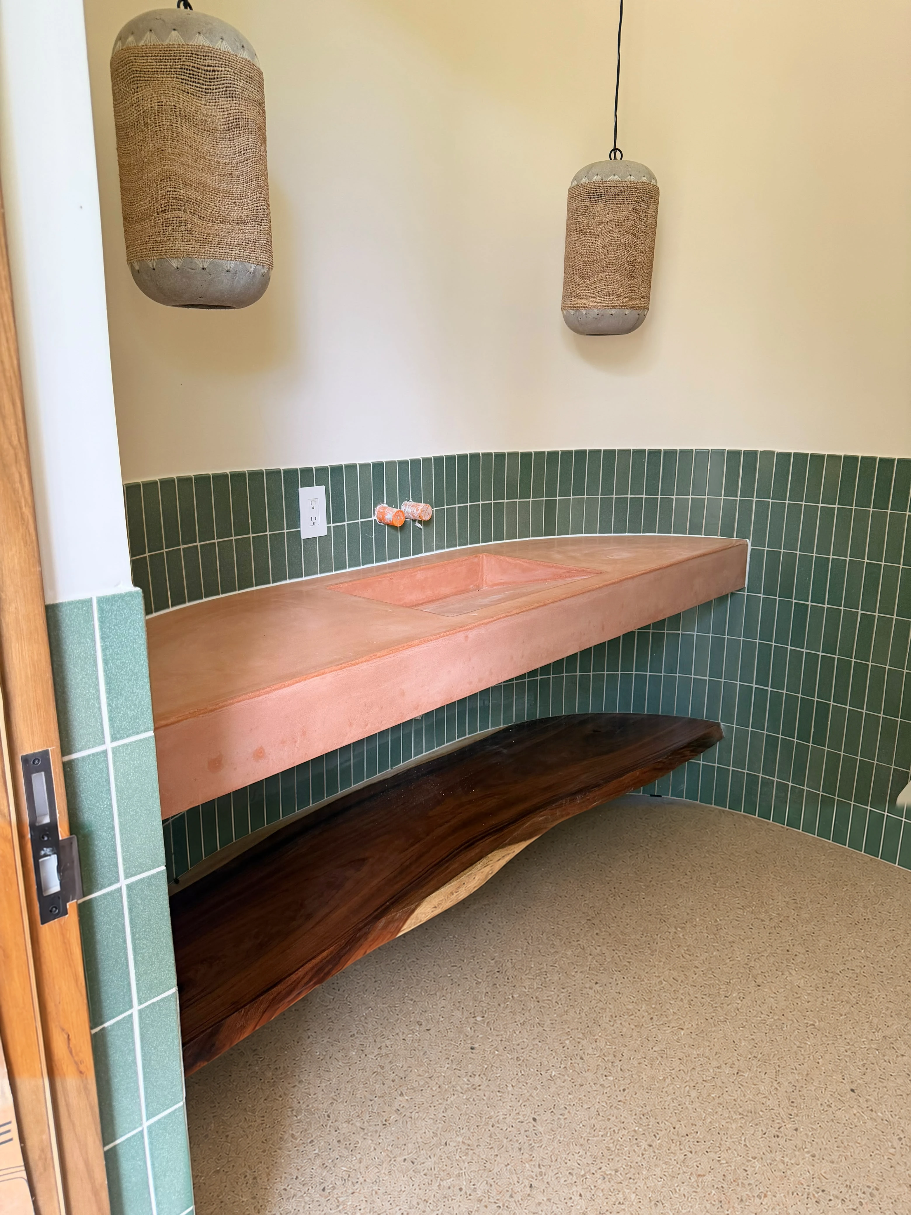 Lavatorio integrado flotante de concreto terracota con repisa de madera natural, en baño con azulejos verdes y lámparas de fibra