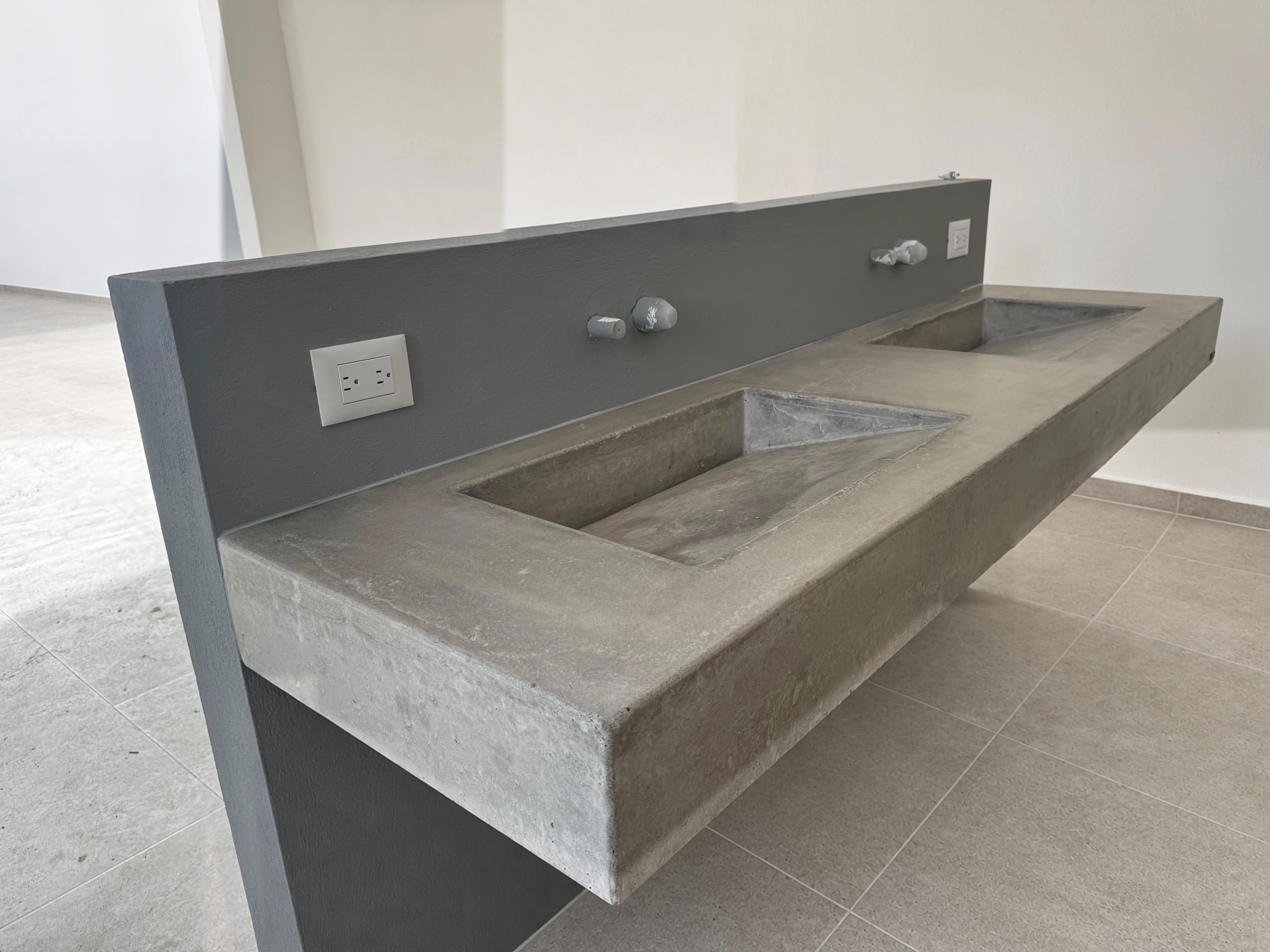 Lavatorio integrado de concreto gris con dos pozas alargadas y respaldo de pared gris, con grifería de pared, en espacio comercial
