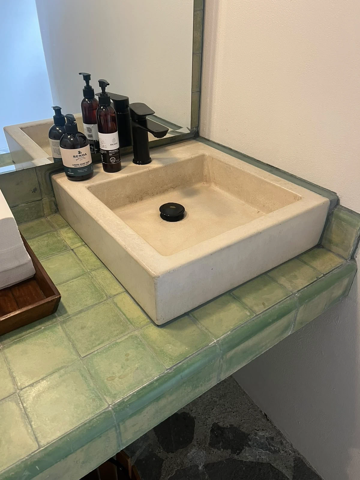 Lavatorio cuadrado de concreto crema con desagüe negro, apoyado sobre encimera de azulejos verdes junto a una grifería negra