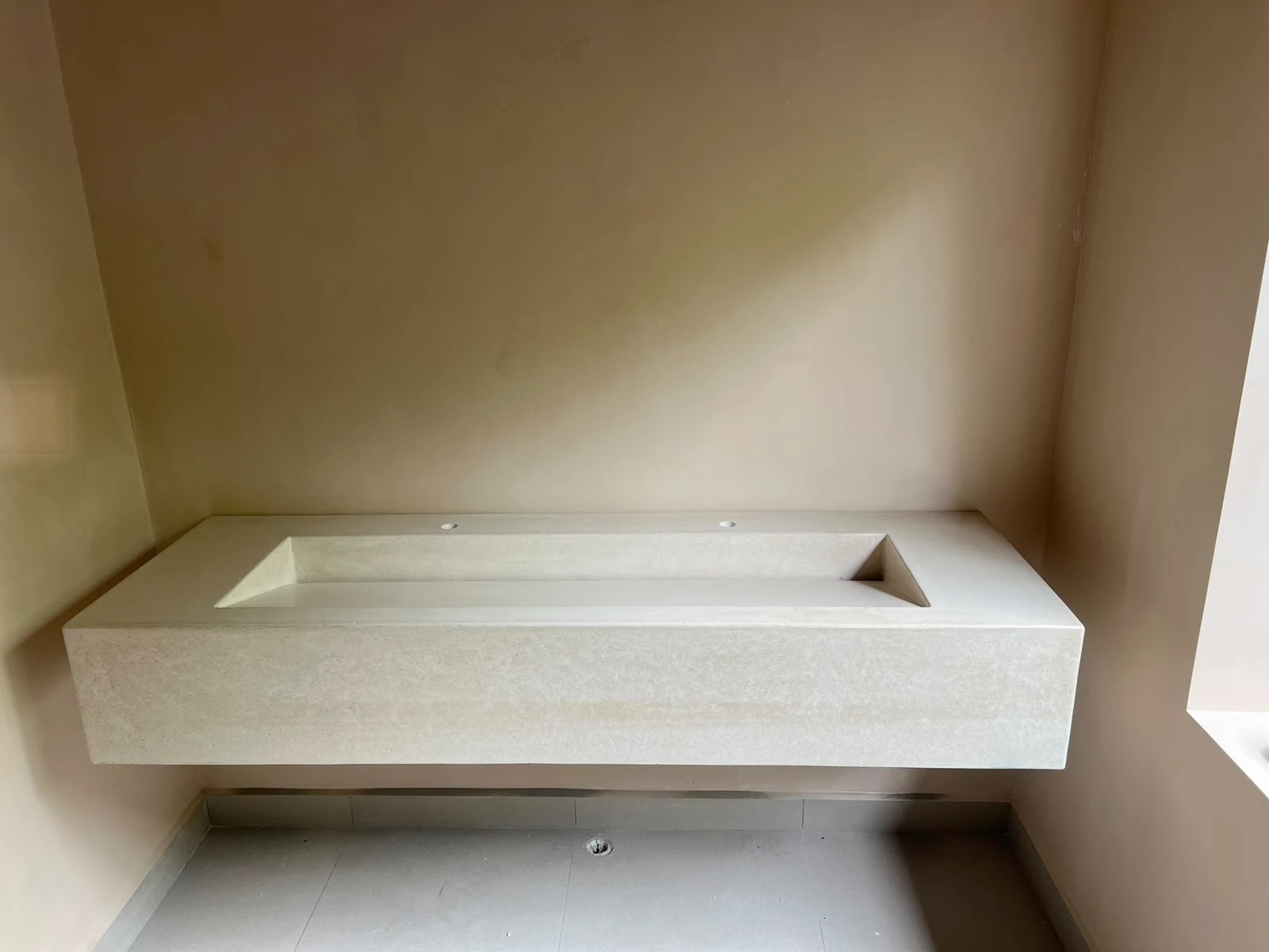 Lavatorio integrado flotante de concreto color arena con poza rectangular alargada, en un nicho de pared de tono claro