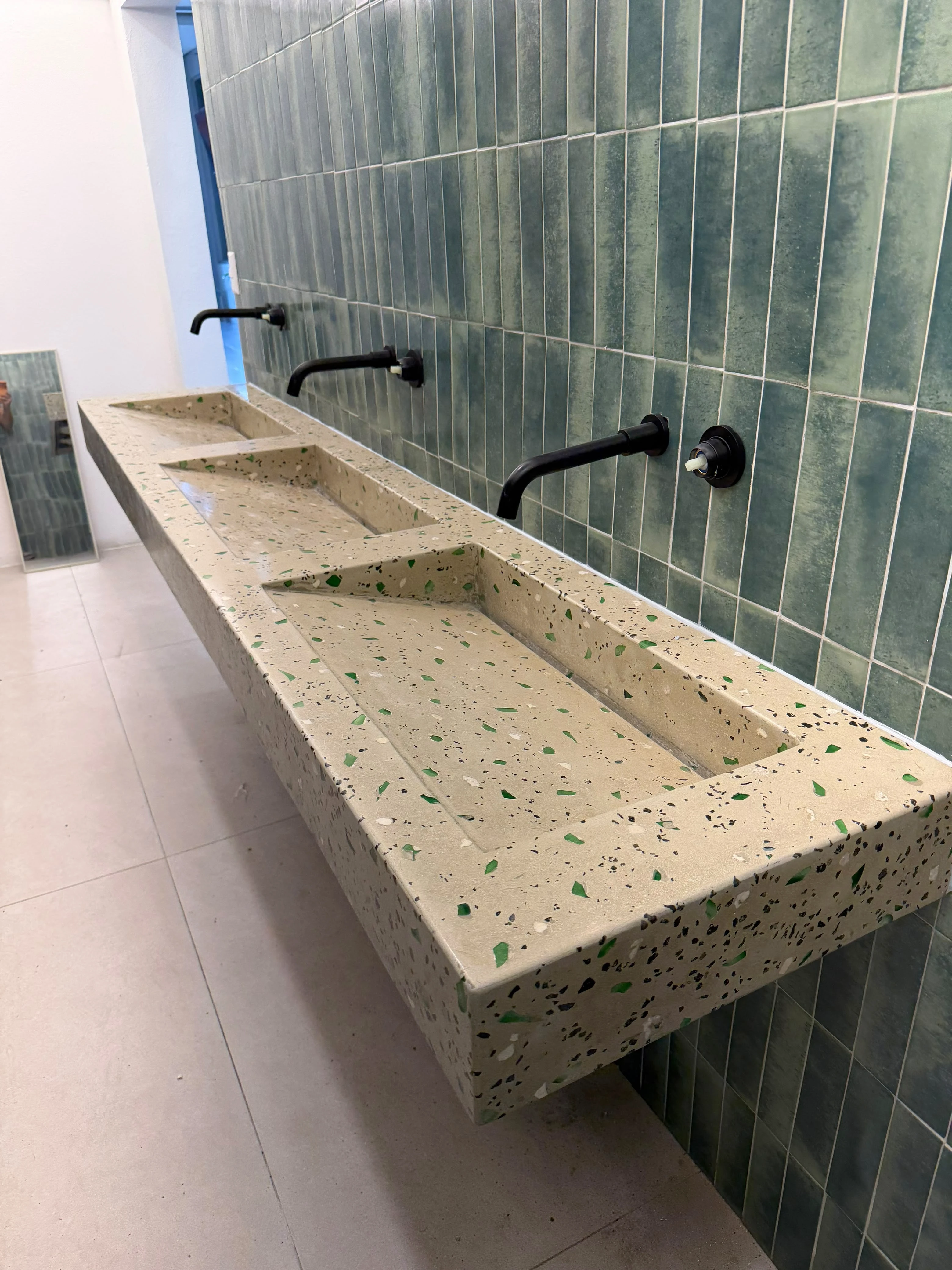 Lavatorio integrado flotante largo de terrazo claro con agregados verdes, tres pozas y grifería negra, sobre pared de azulejos verdes