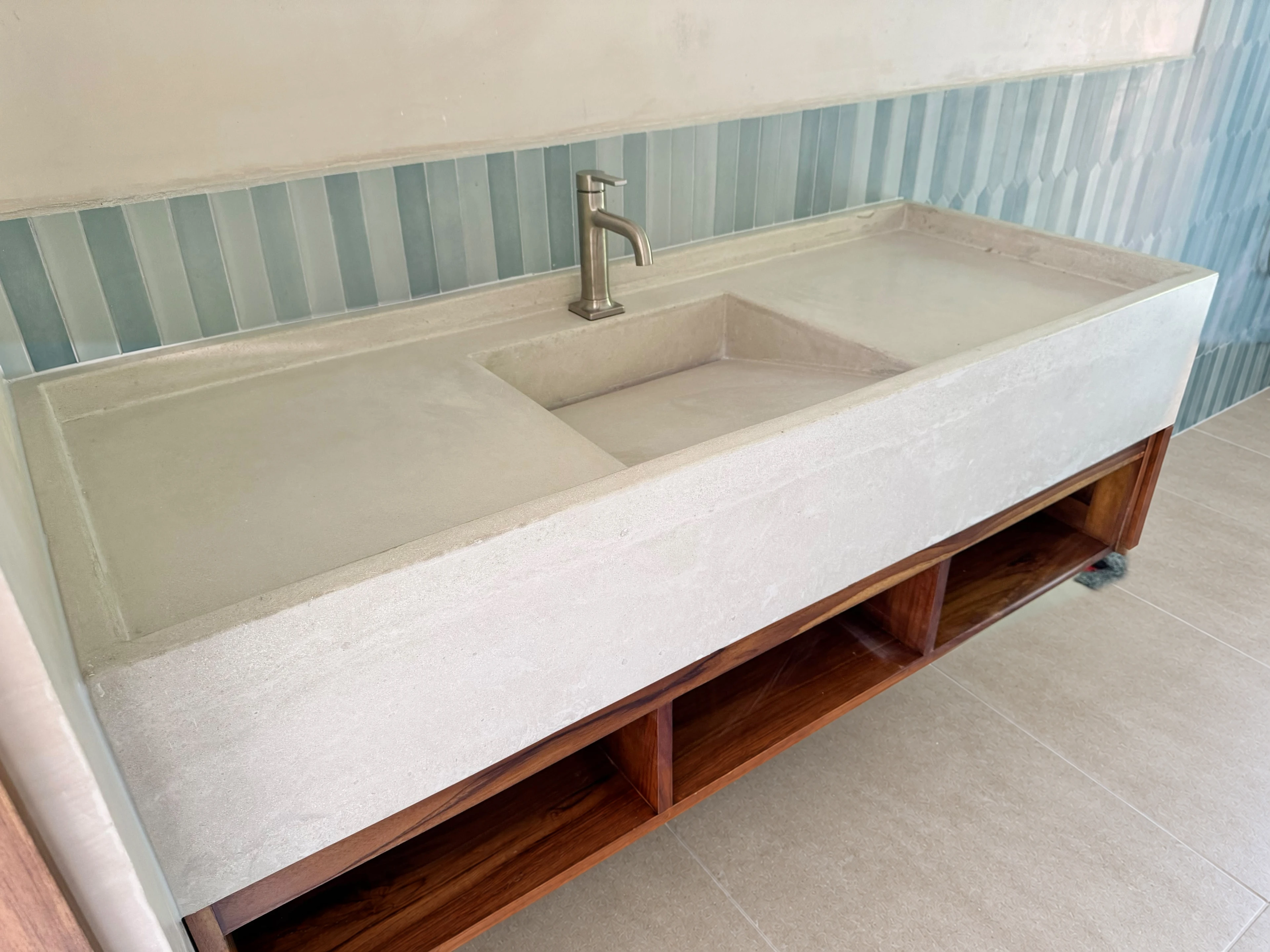 Lavatorio integrado largo de concreto claro con poza inclinada y grifería cromada, sobre mueble de madera abierto y azulejos celestes