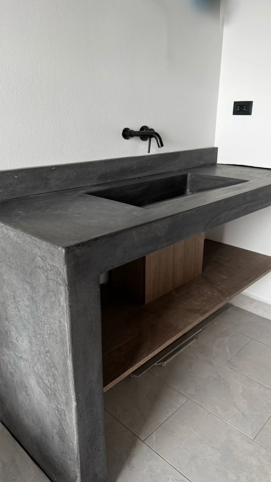 Lavatorio integrado de concreto gris oscuro con poza alargada, grifería negra de pared y mueble con repisa de madera