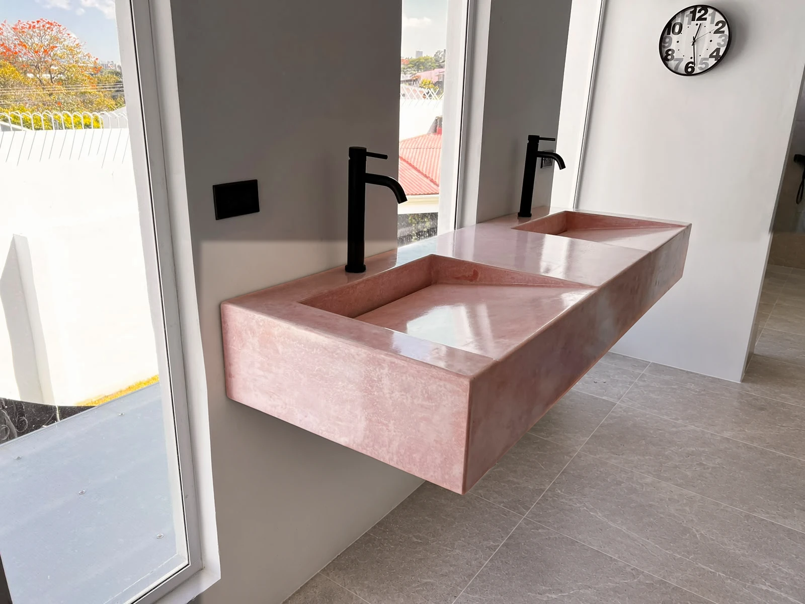 Lavatorio integrado flotante doble de concreto rosado pulido con grifería negra, junto a ventanales luminosos