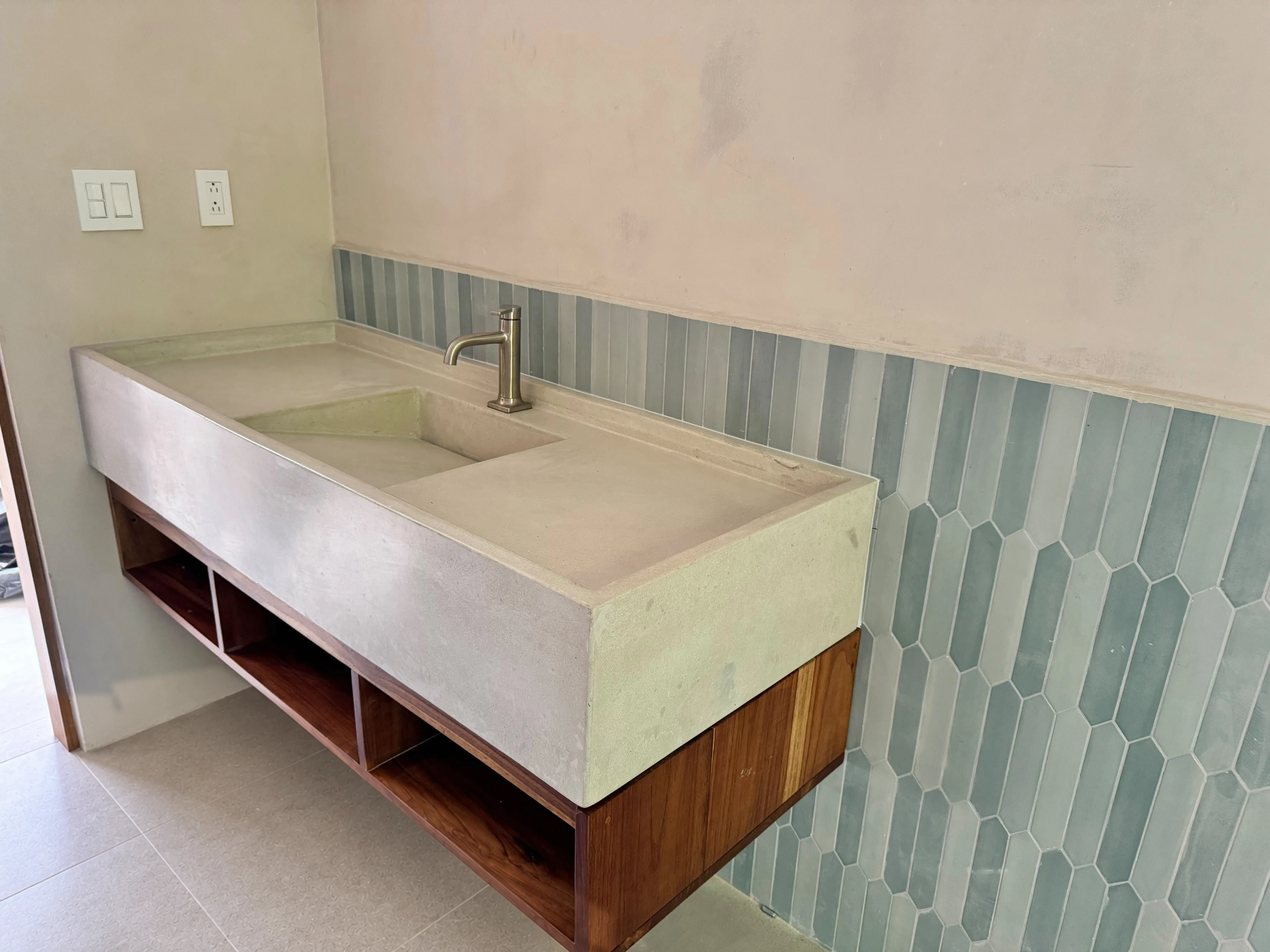 Lavatorio integrado de concreto claro con poza alargada y grifería cromada, sobre mueble de madera flotante y azulejos celestes