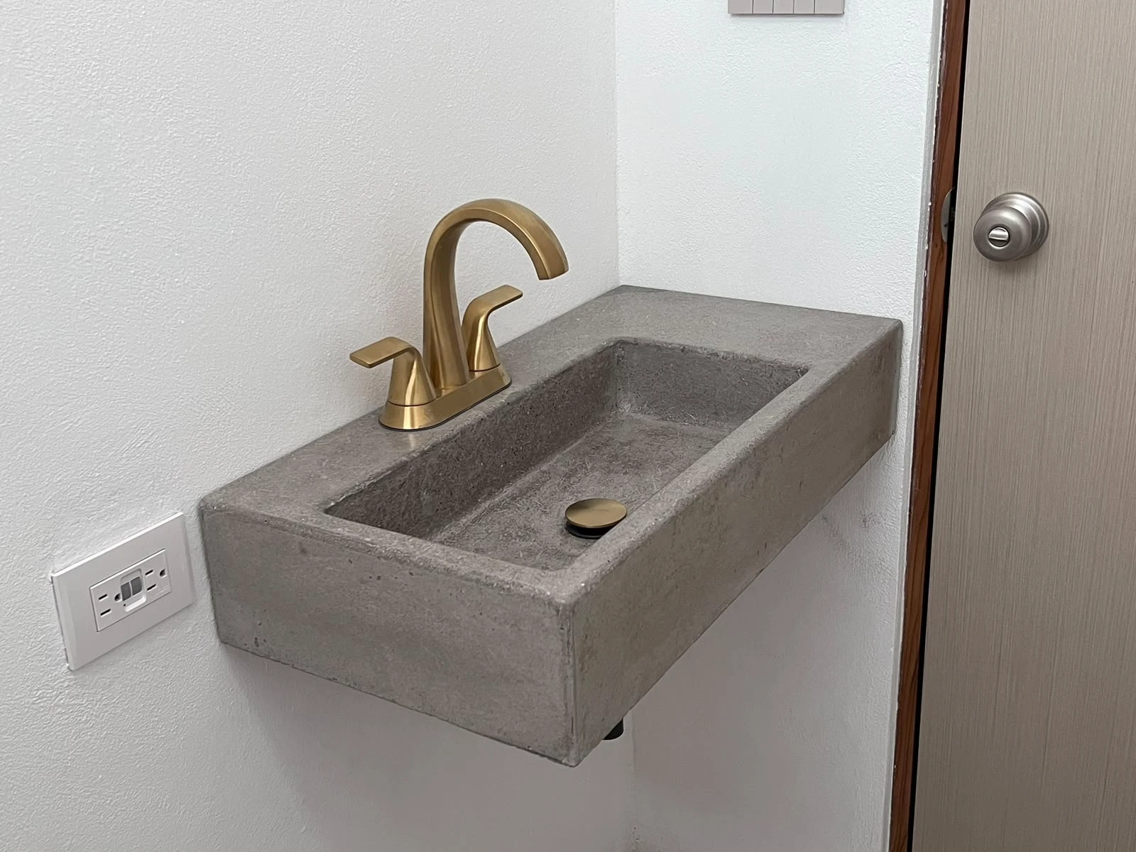 Lavatorio integrado pequeño de concreto gris con poza rectangular y grifería dorada, fijado a una pared blanca