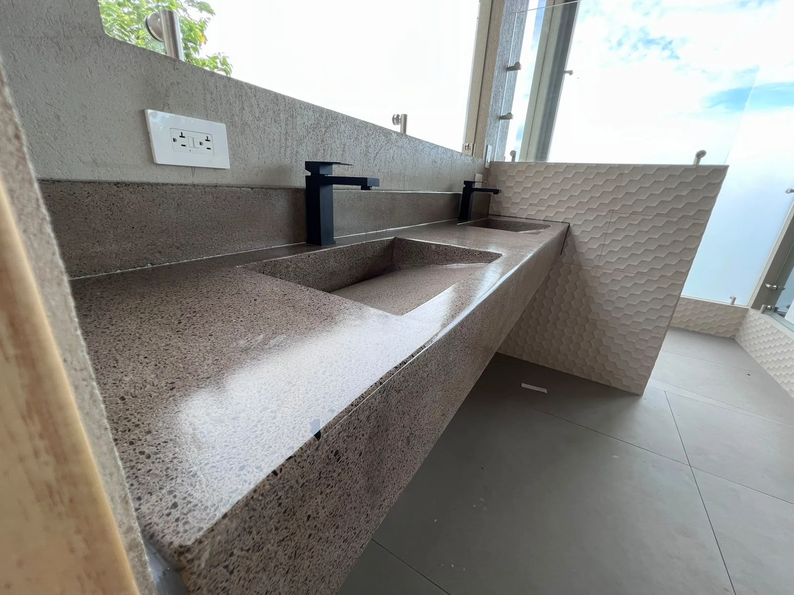 Lavatorio integrado largo de concreto rosado pardo con grifería negra y pared de relieve, junto a ventanal