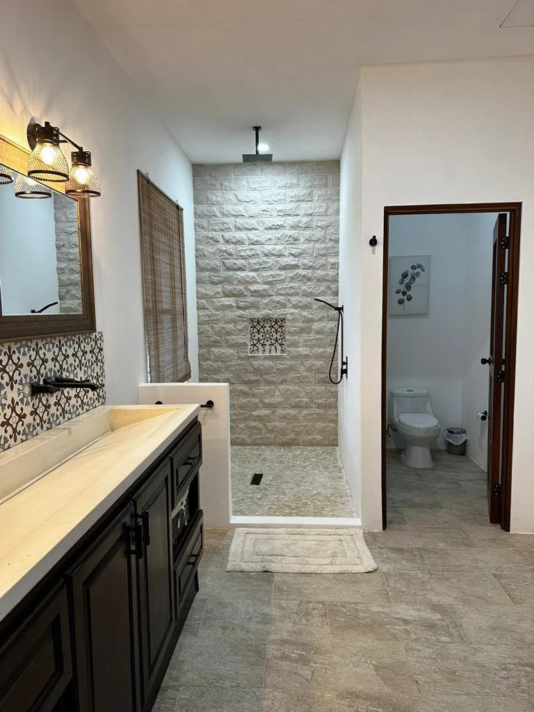 Lavatorio integrado largo de concreto crema con canal sobre mueble de madera oscura, en baño con ducha de piedra al fondo