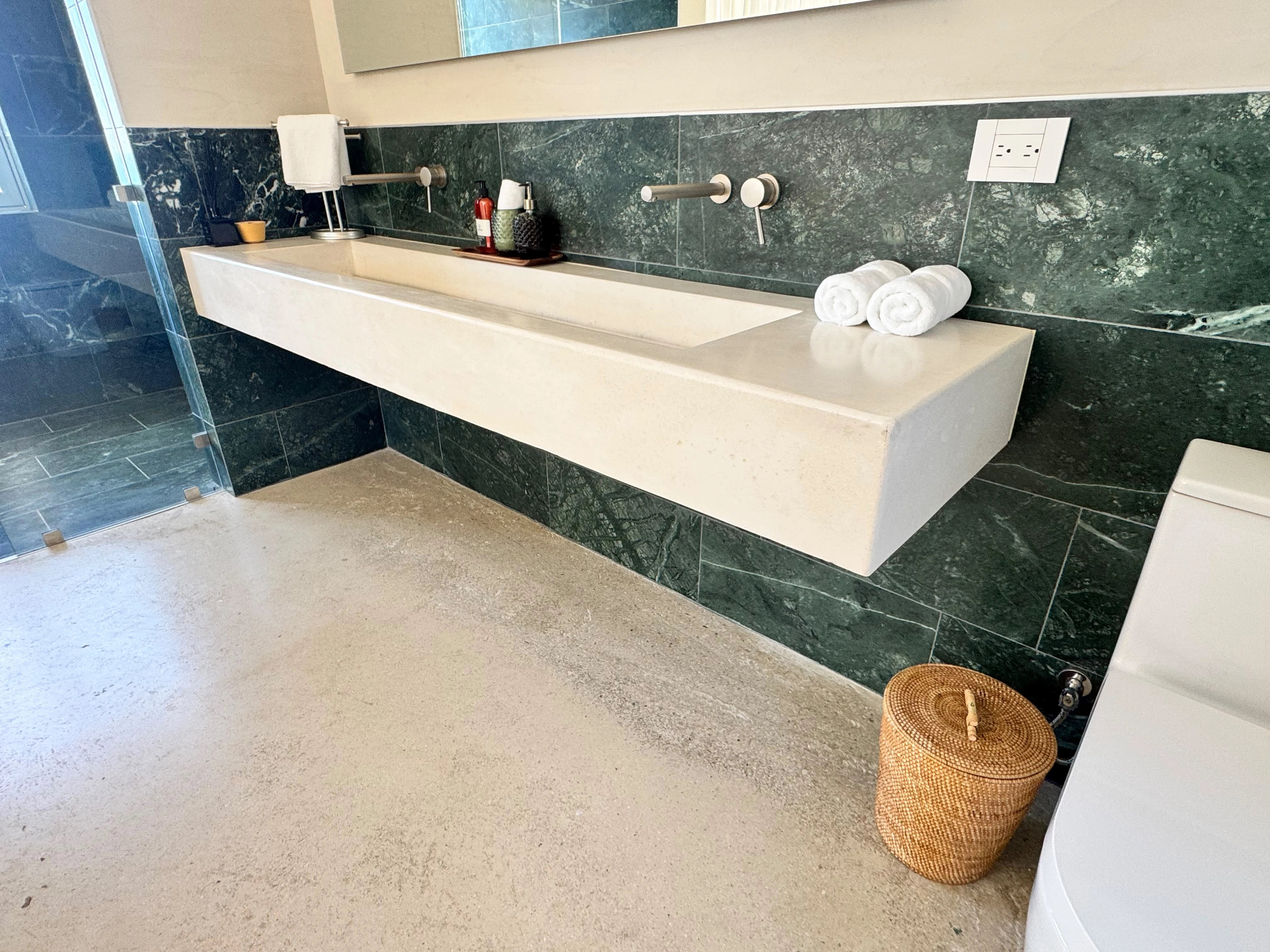 Lavatorio integrado flotante de concreto crema con canal alargado y grifería de pared, sobre pared de mármol verde oscuro