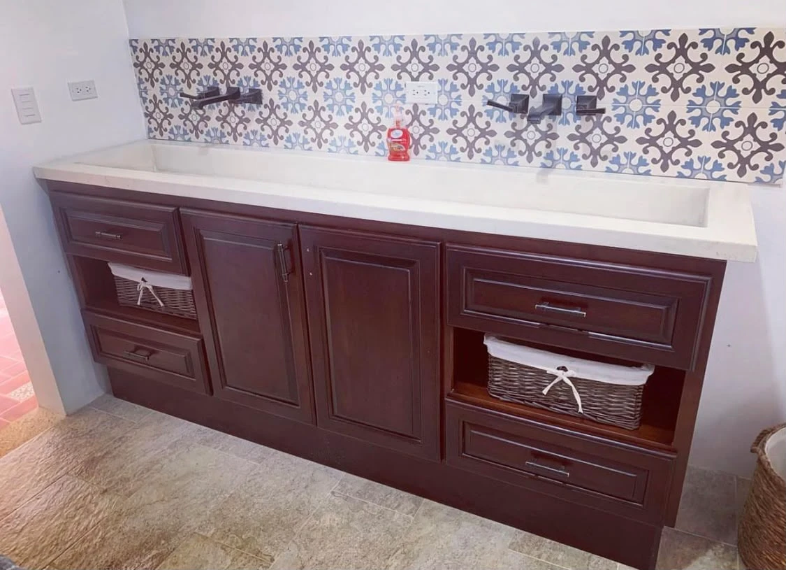 Lavatorio integrado largo de concreto claro con canal poco profundo, sobre mueble de madera rojiza y frente de azulejos decorados