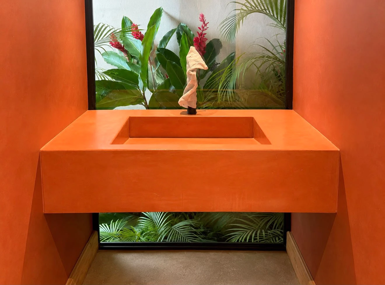Lavatorio integrado flotante de concreto naranja intenso con poza rectangular, bajo una ventana con plantas tropicales