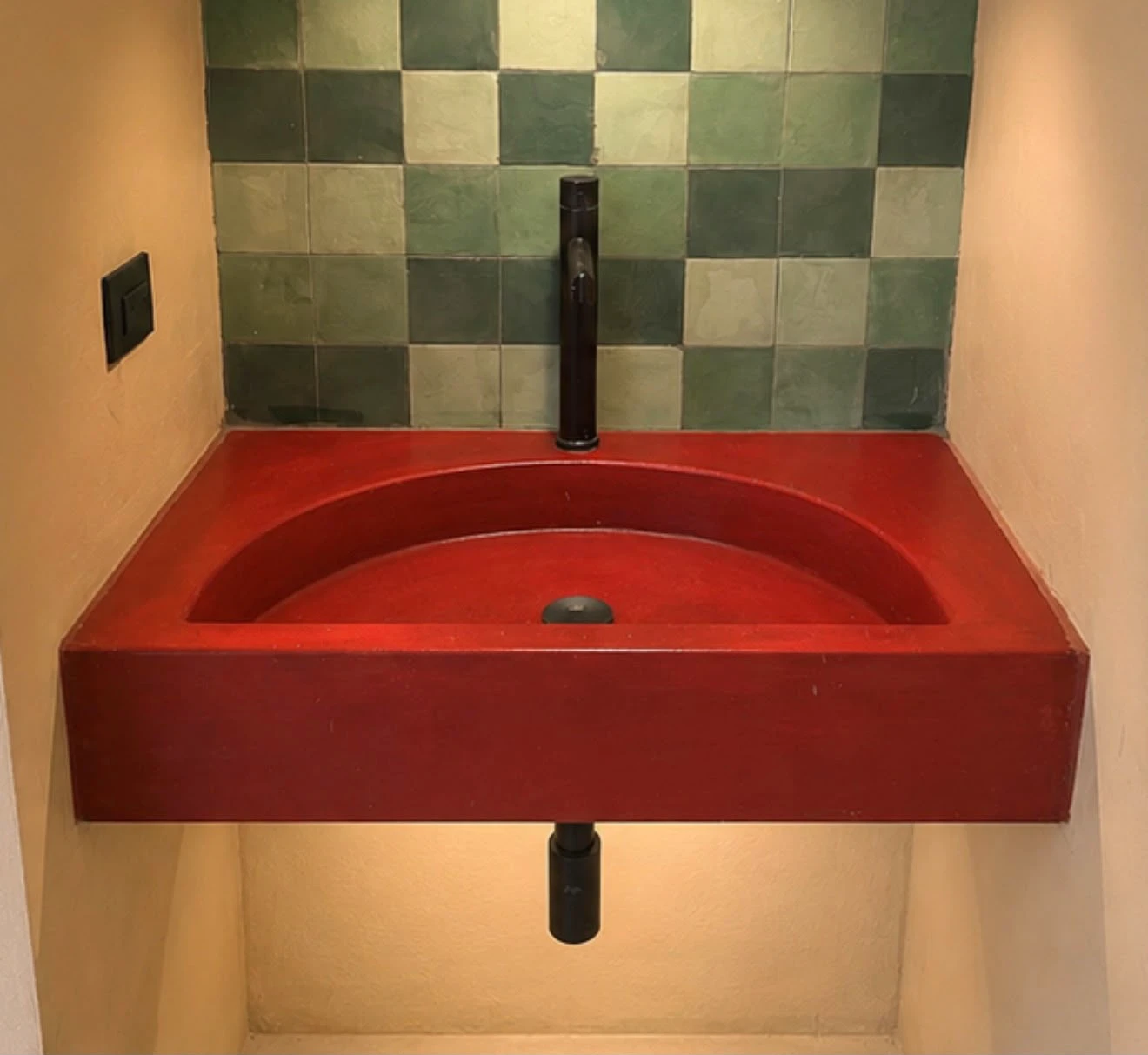 Lavatorio integrado flotante de concreto rojo con poza semicircular, grifería negra y pared de azulejos verdes