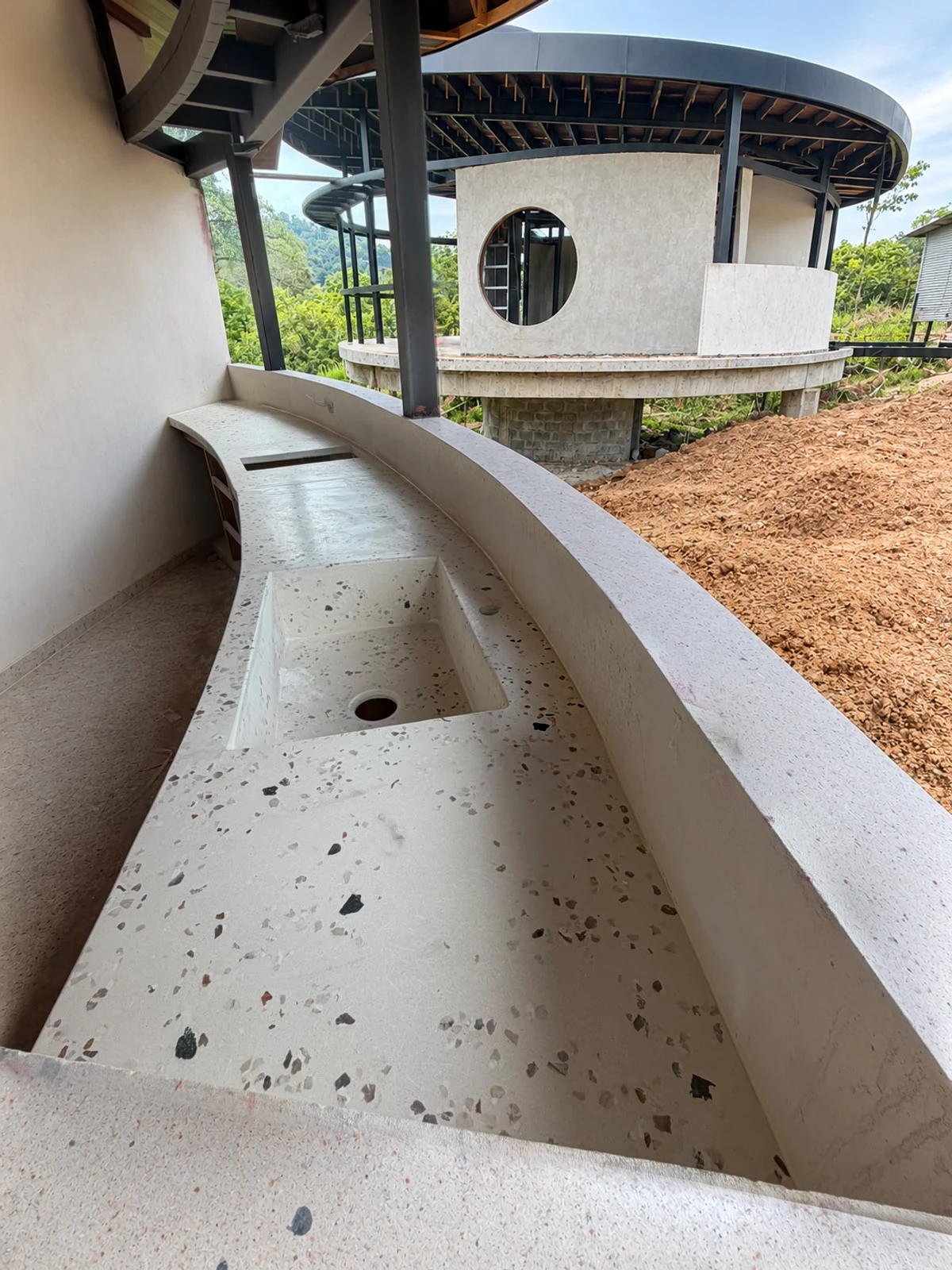 Lavatorio integrado curvo de terrazo claro con agregados oscuros y poza alargada, en obra al aire libre con vista al terreno