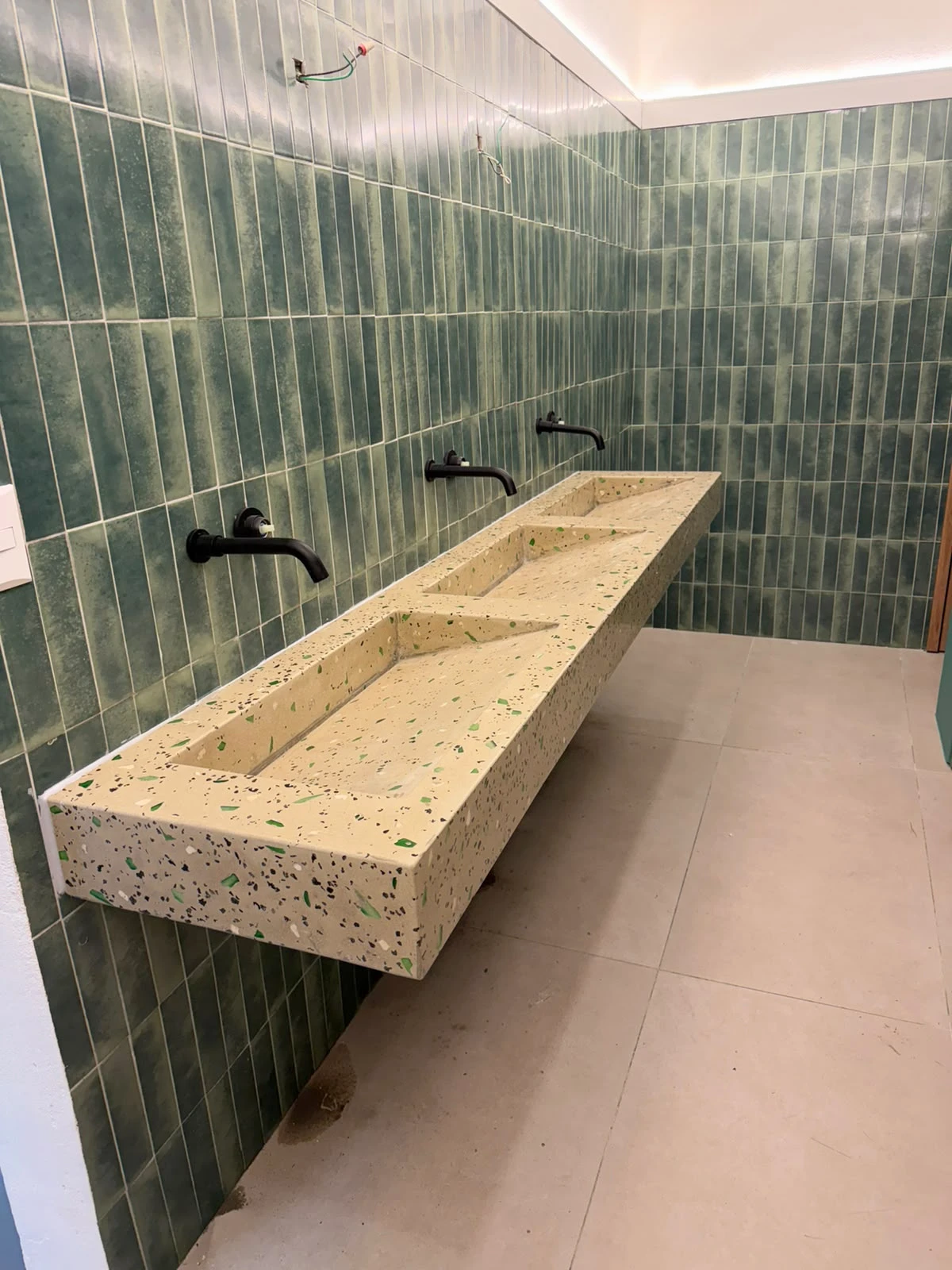 Lavatorio integrado largo de terrazo claro con agregados de colores y dos pozas, sobre pared de azulejos verdes y grifería negra