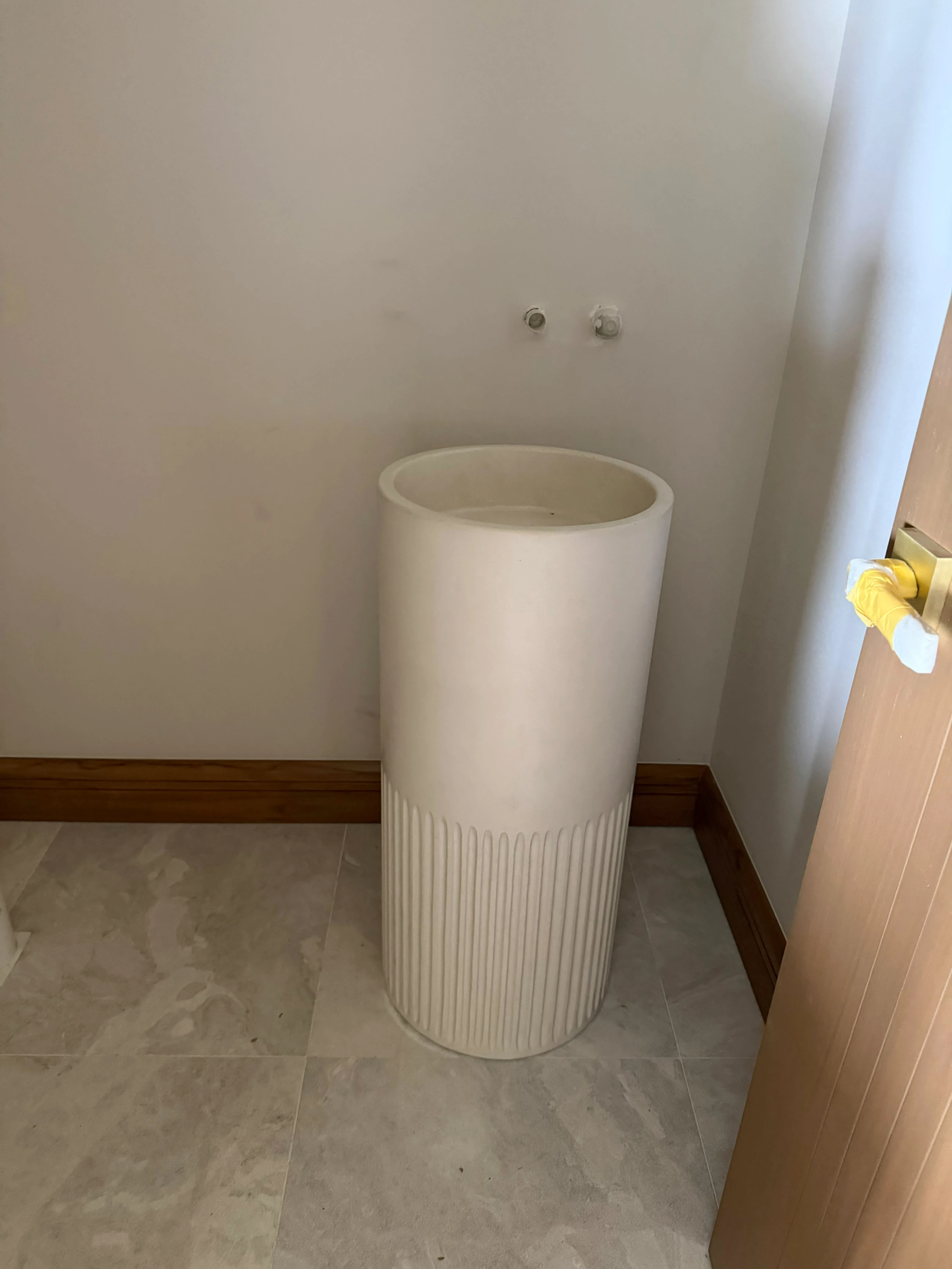 Lavatorio de pedestal cilíndrico de concreto blanco con base acanalada, en baño con grifería de pared y piso de losa