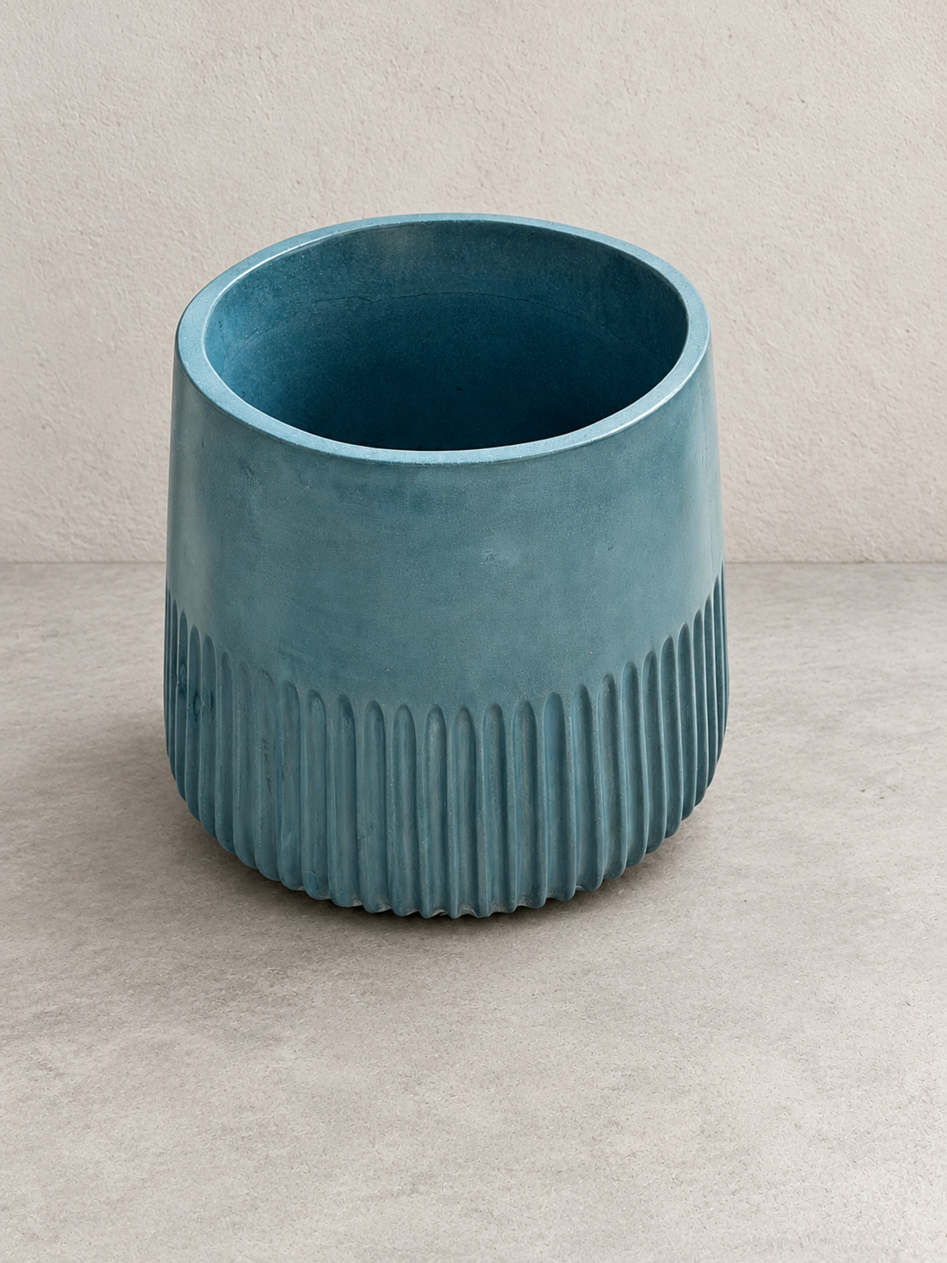 Lavatorio de pedestal cilíndrico de concreto turquesa con base acanalada y boca ancha, sobre piso claro frente a pared lisa