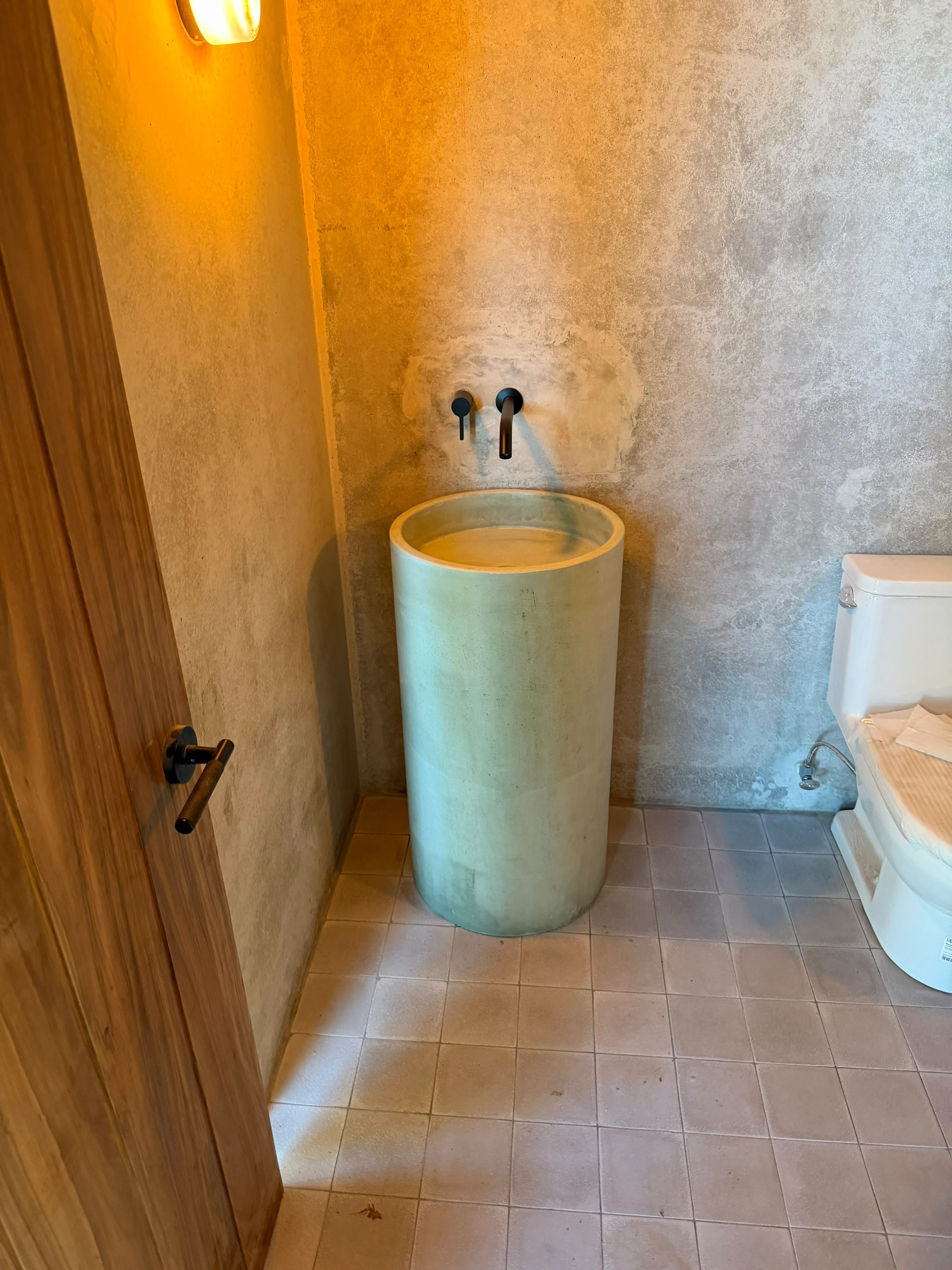 Lavatorio de pedestal cilíndrico de concreto verde menta con grifería de pared negra, en baño con paredes de textura rústica