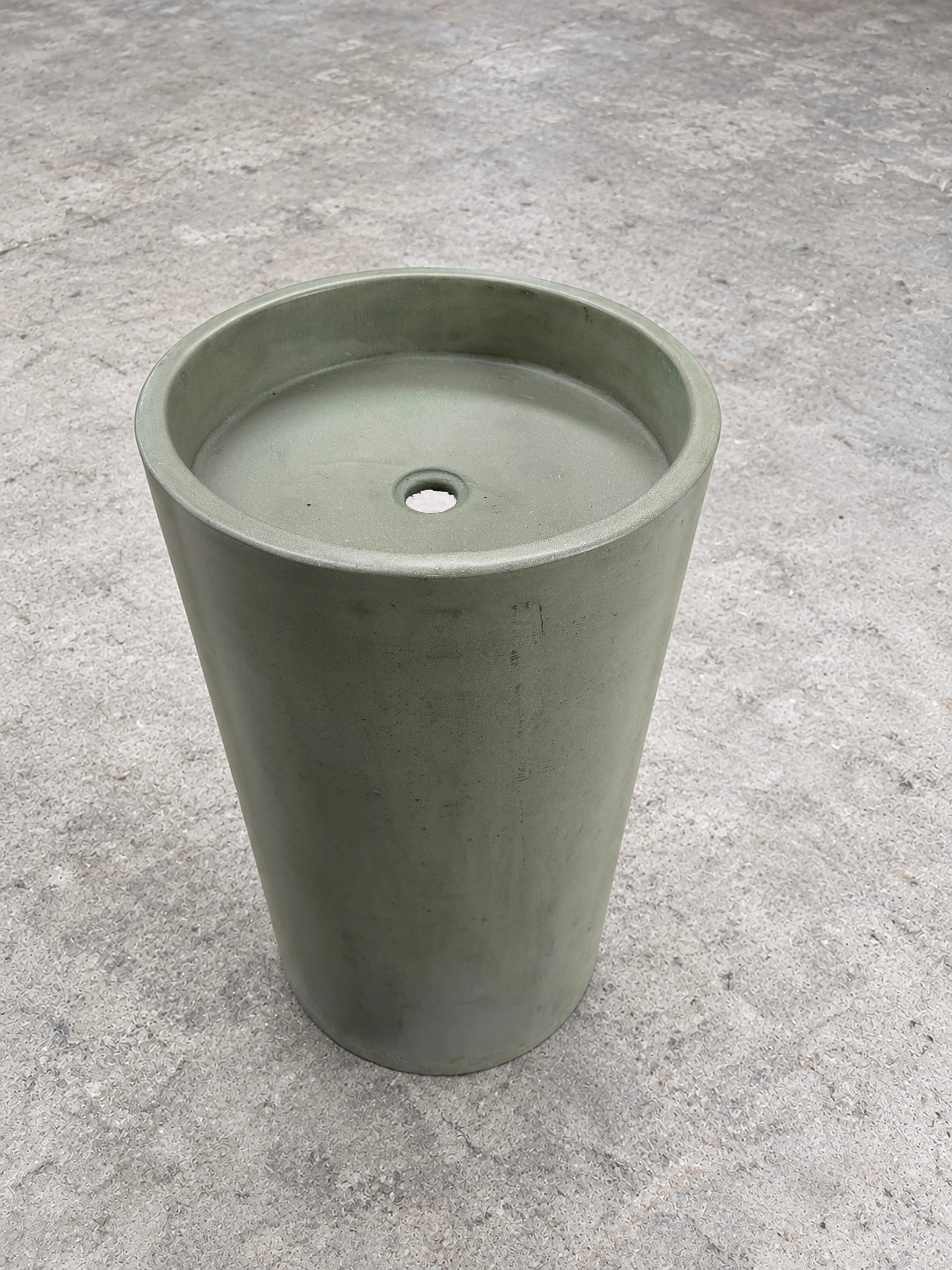 Lavatorio de pedestal cónico de concreto verde oliva con poza circular y desagüe central, sobre piso gris de cemento pulido