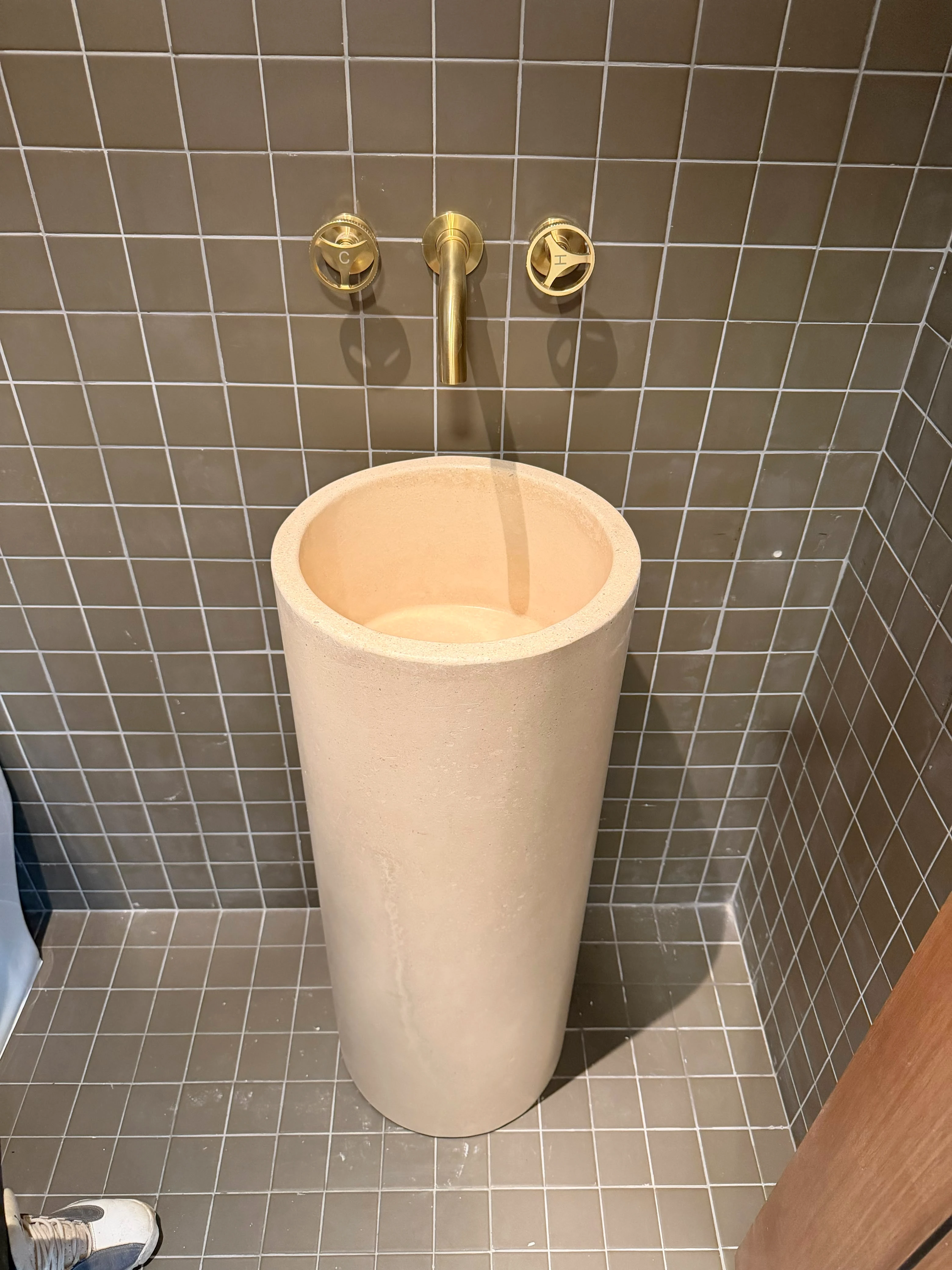 Lavatorio de pedestal cónico de concreto color crema con grifería dorada de pared, en baño con azulejos verde oliva