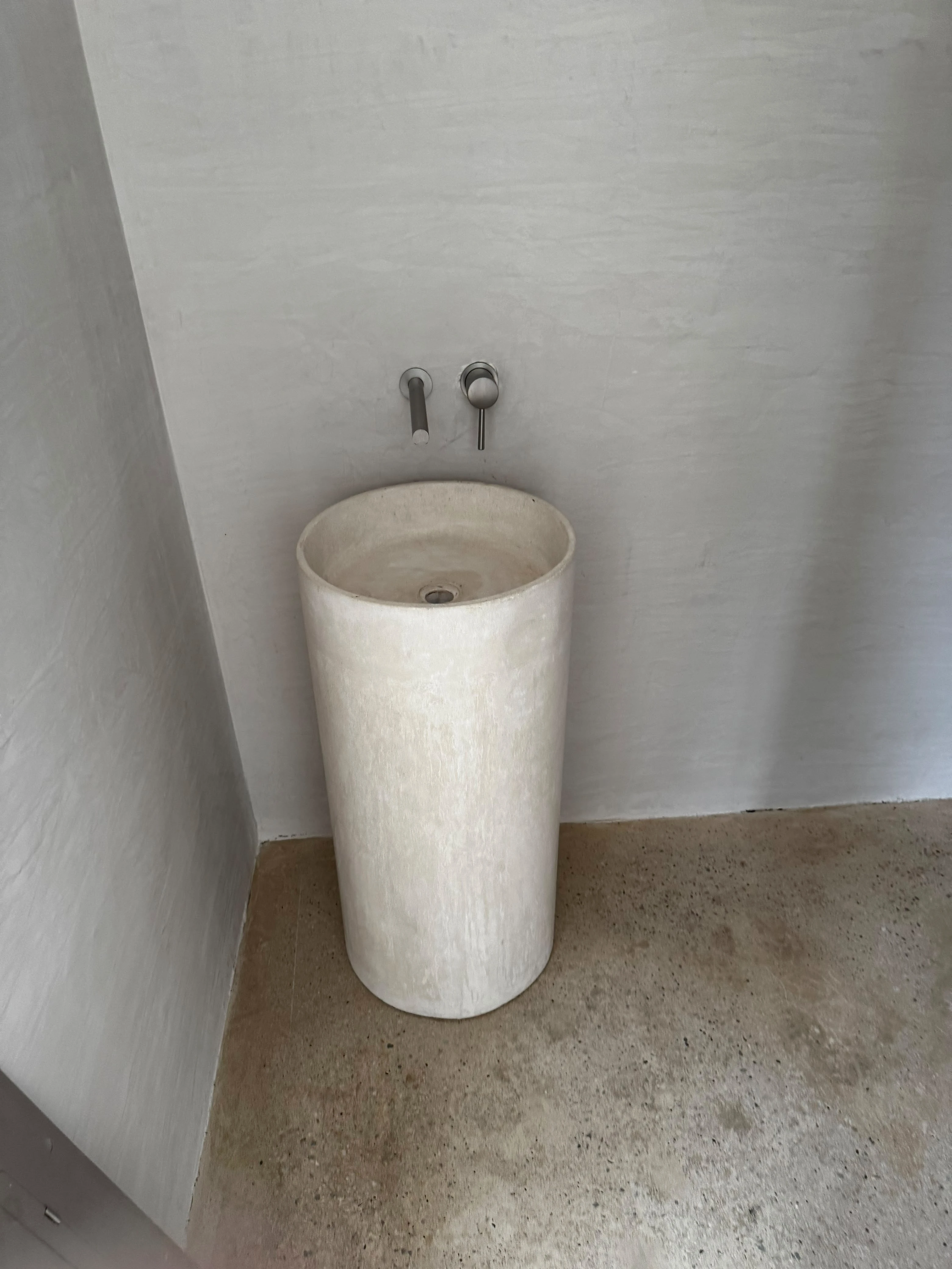 Lavatorio de pedestal cilíndrico de concreto claro con grifería de pared, en esquina de baño con paredes de textura rústica