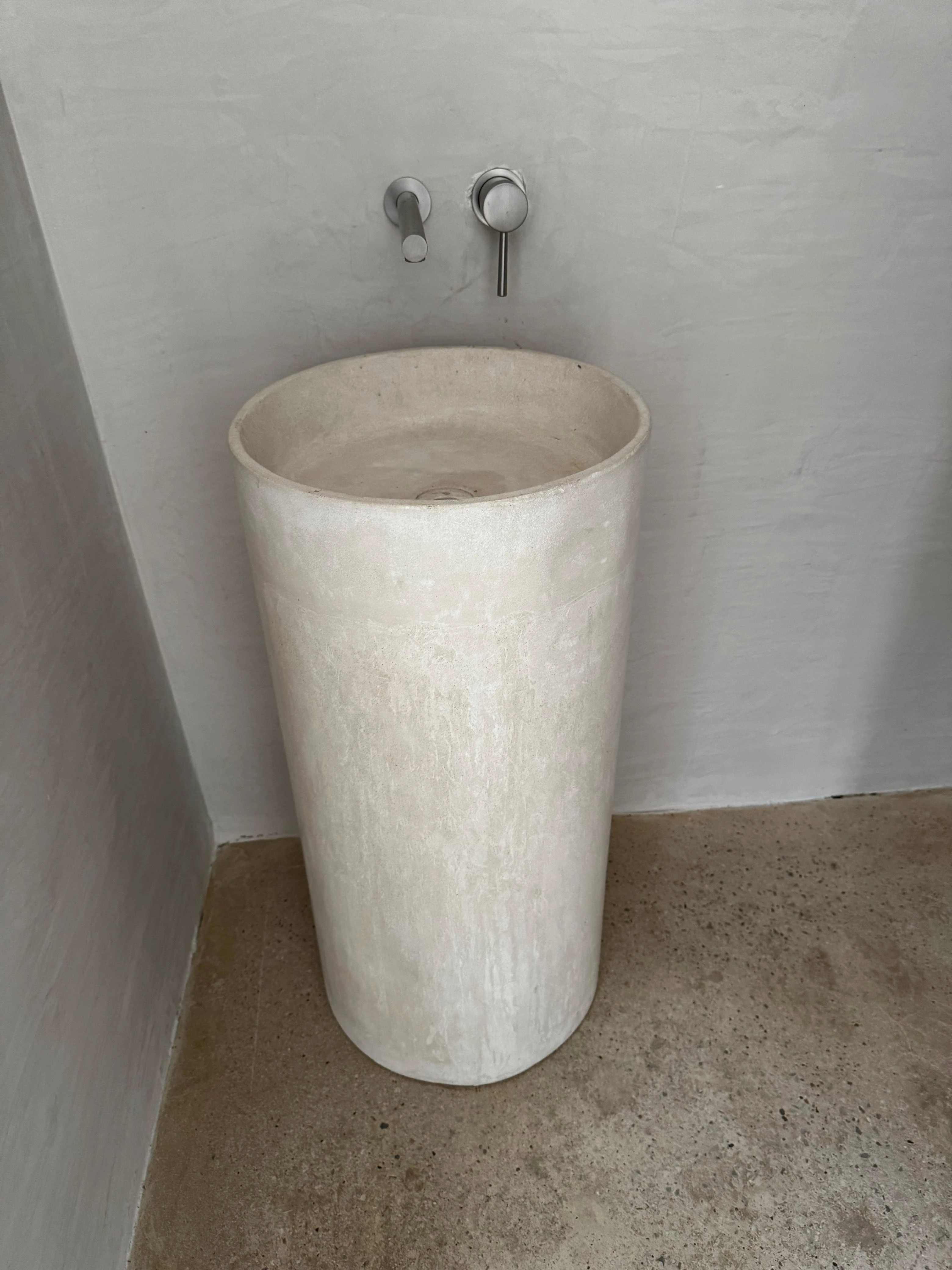 Lavatorio de pedestal cilíndrico de concreto gris claro con grifería de pared cromada, en baño con paredes blancas texturizadas