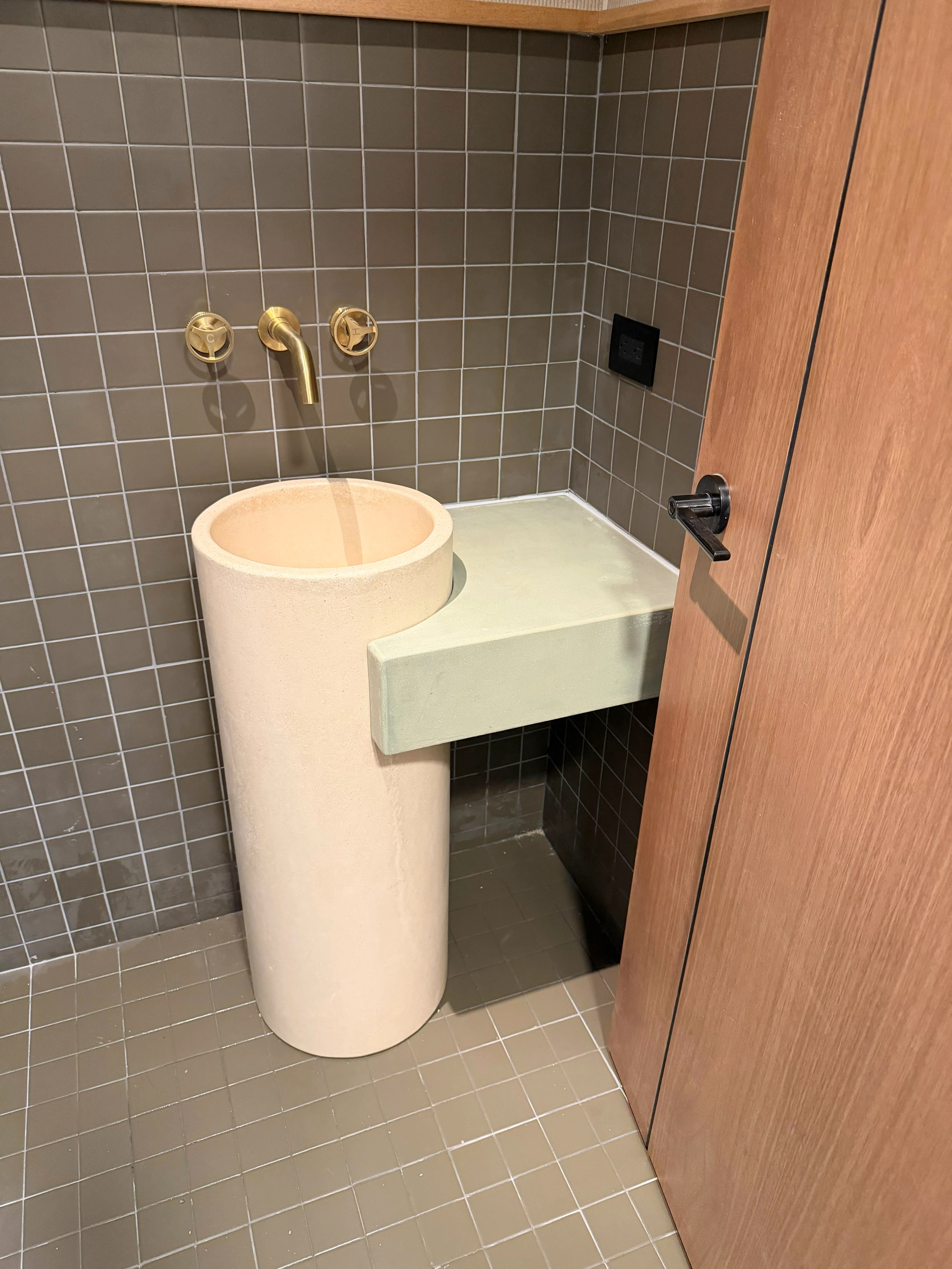 Lavatorio de pedestal cilíndrico de concreto claro con repisa lateral verde, grifería dorada y azulejos oscuros en baño