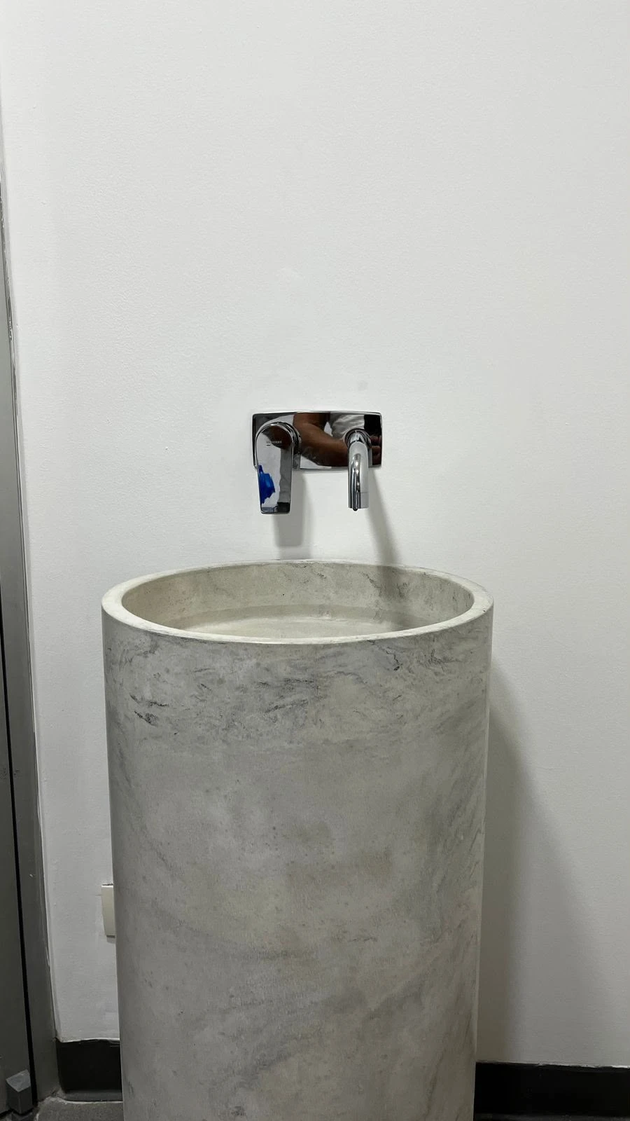 Lavatorio de pedestal cilíndrico de concreto gris con vetas, con grifería cromada de pared, en baño de paredes blancas