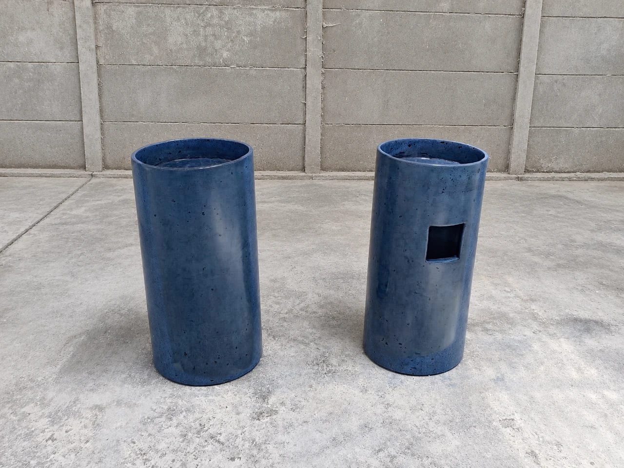 Dos lavatorios de pedestal cilíndricos de concreto azul intenso con una abertura cuadrada, sobre piso de cemento y muro de bloques