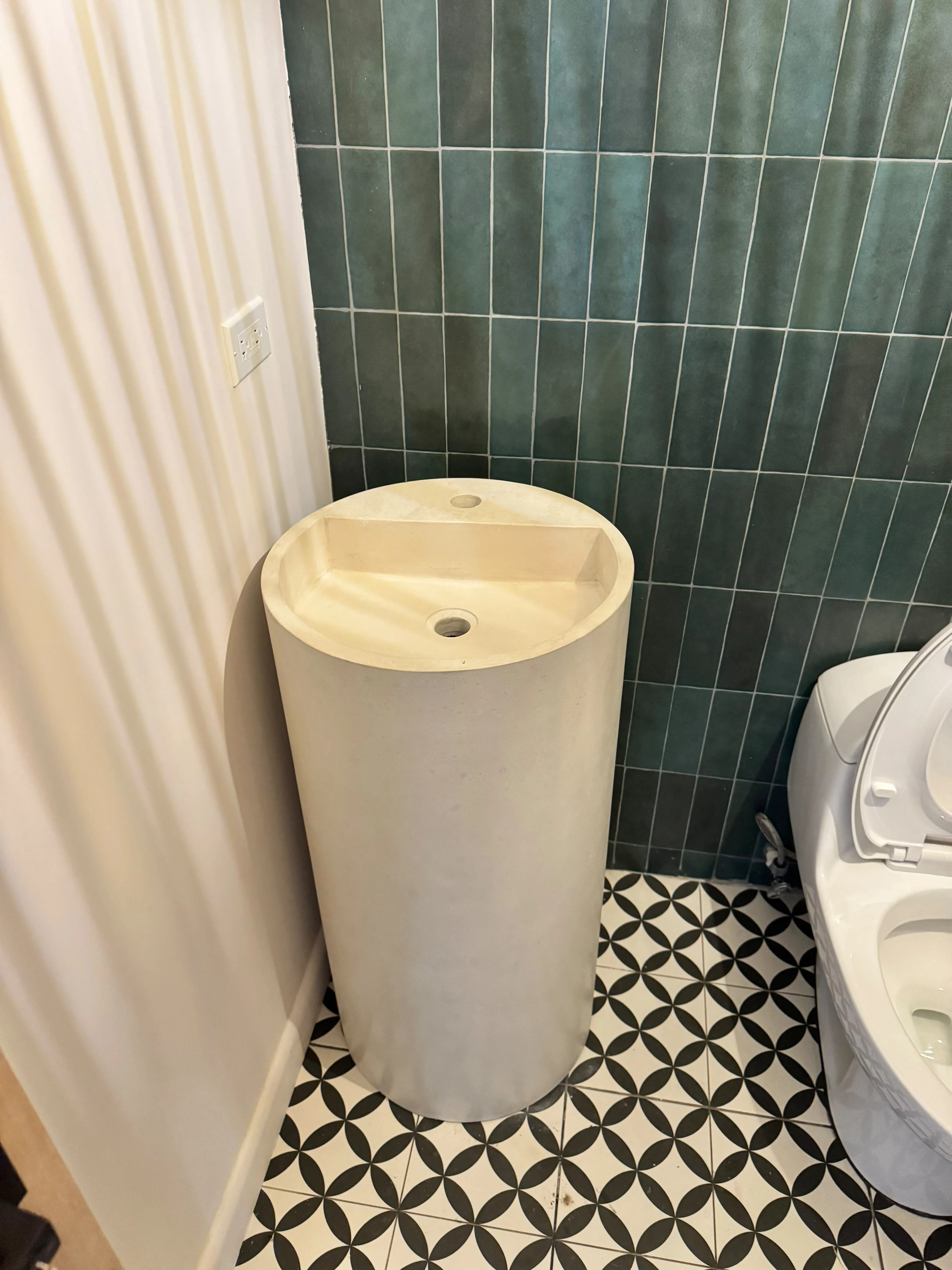 Lavatorio de pedestal cilíndrico de concreto color crema, en baño con azulejos verdes alargados y piso de mosaico