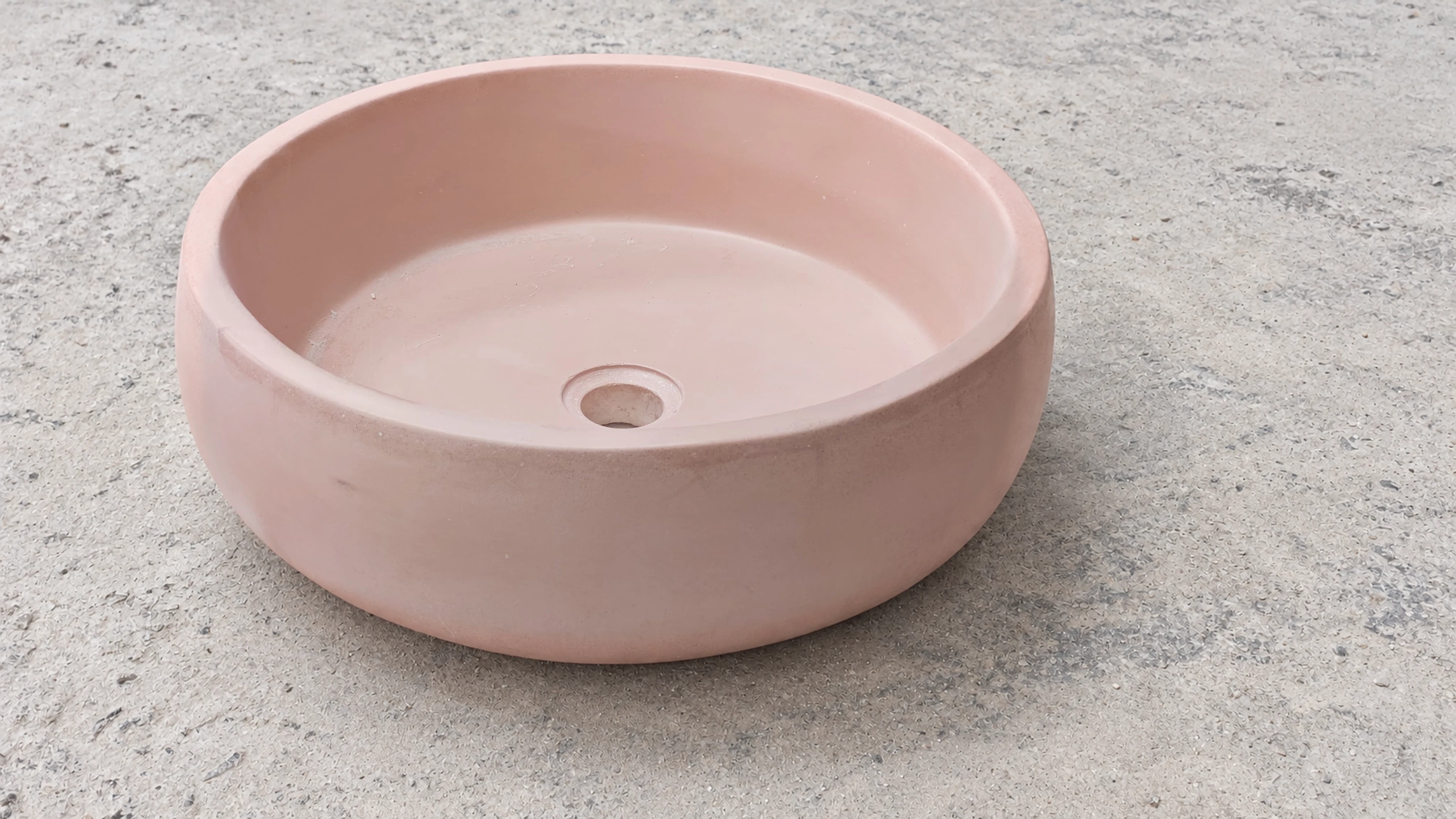 Lavatorio de sobreponer redondo de concreto rosado pálido con borde grueso y curvo, sobre superficie gris texturizada