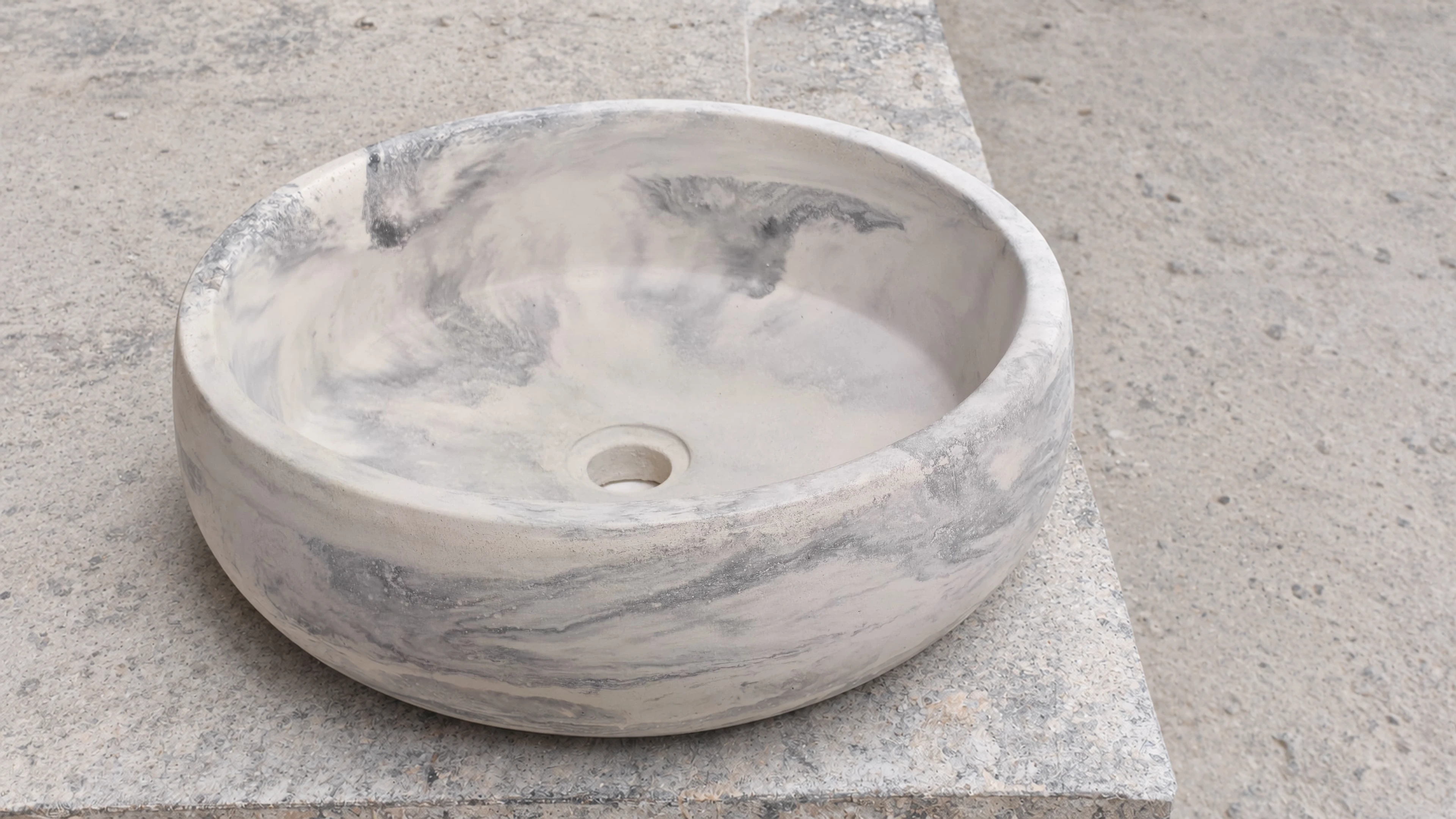 Lavatorio de sobreponer redondo de concreto con vetas grises y blancas tipo mármol, sobre superficie de piedra clara