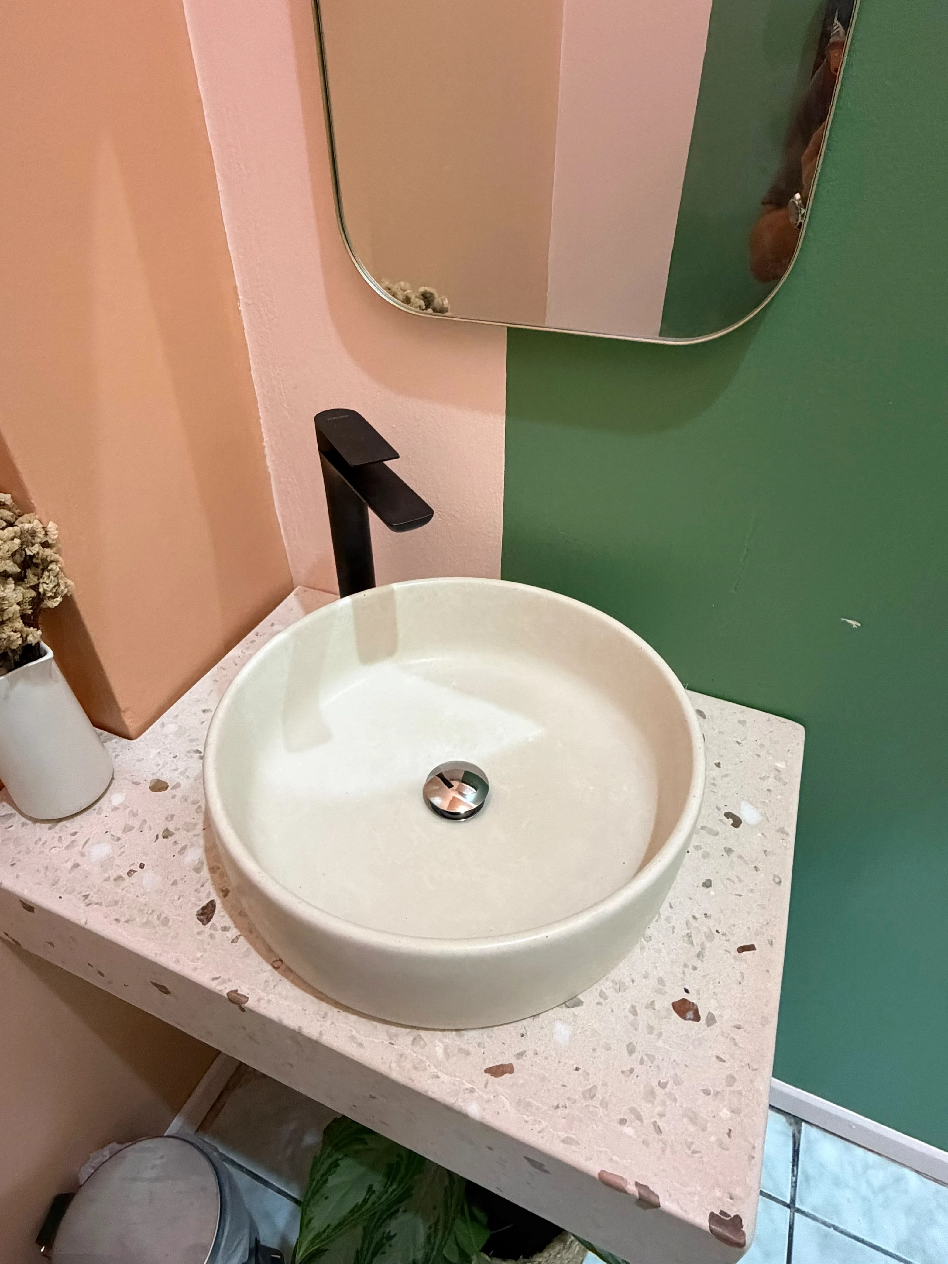 Lavatorio de sobreponer redondo de concreto blanco crema sobre encimera de terrazo rosado, grifería negra alta y pared verde y rosada