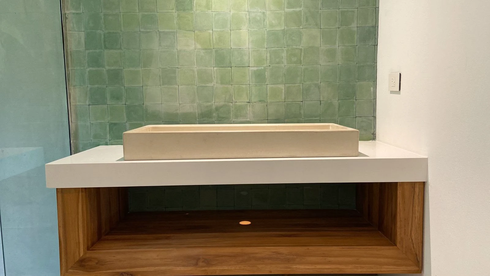 Lavatorio de sobreponer rectangular de concreto color arena sobre encimera blanca flotante con repisa de madera y azulejos verdes