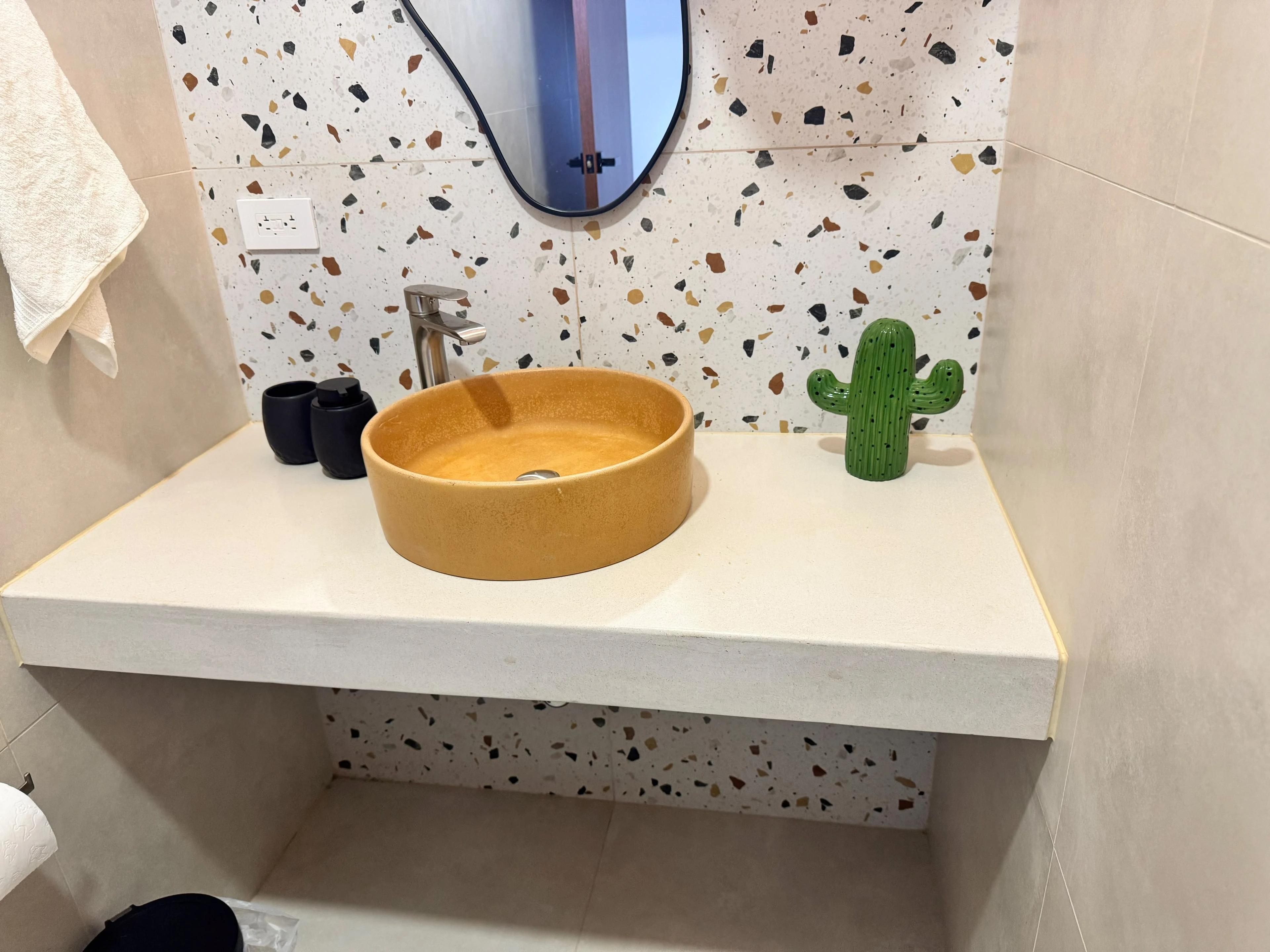 Lavatorio de sobreponer redondo de concreto amarillo mostaza sobre encimera blanca, con grifería cromada y pared de terrazo
