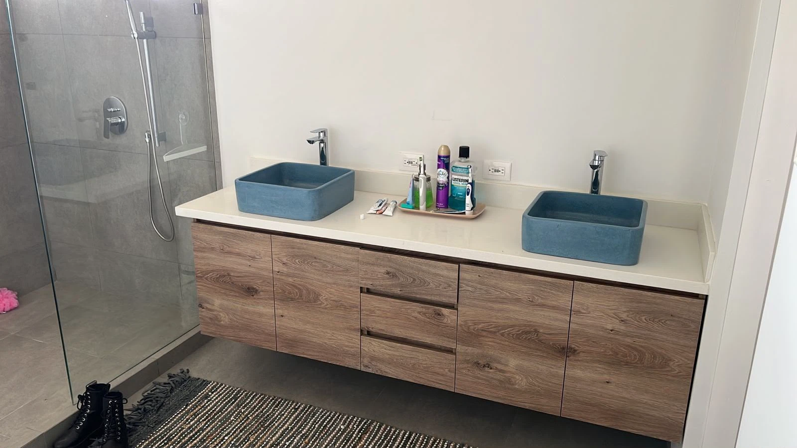 Dos lavatorios de sobreponer rectangulares de concreto azul sobre encimera blanca y mueble de madera, en baño con ducha de vidrio