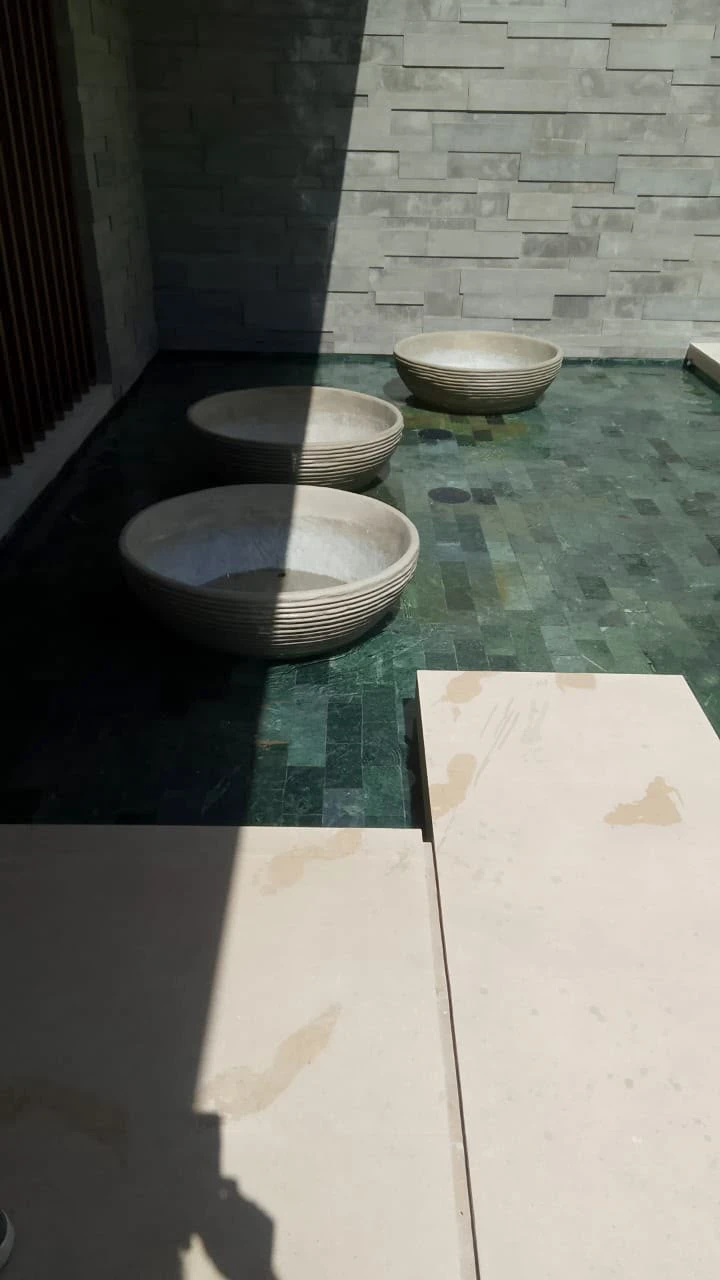 Piezas redondas de concreto claro con textura acanalada, colocadas sobre piso de azulejo verde en un patio con muro de piedra