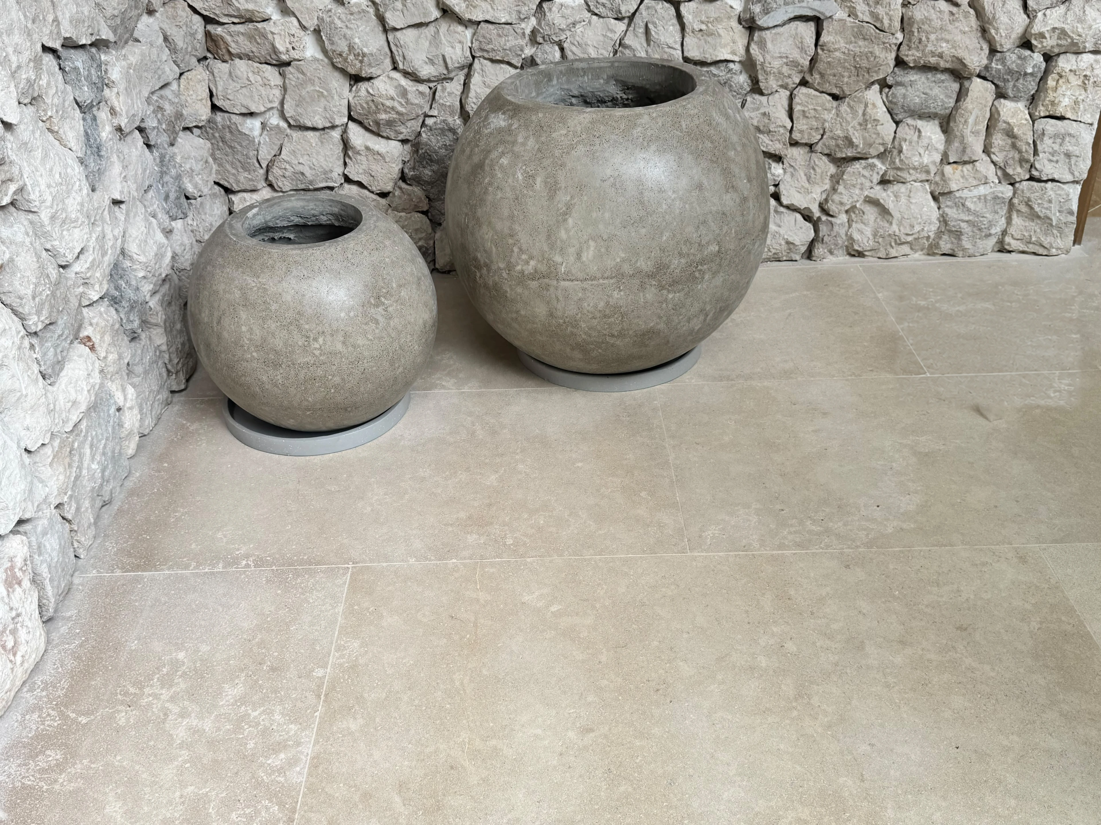 Dos maceteros esféricos de concreto gris de distinto tamaño, sobre piso claro y junto a una pared de piedra natural