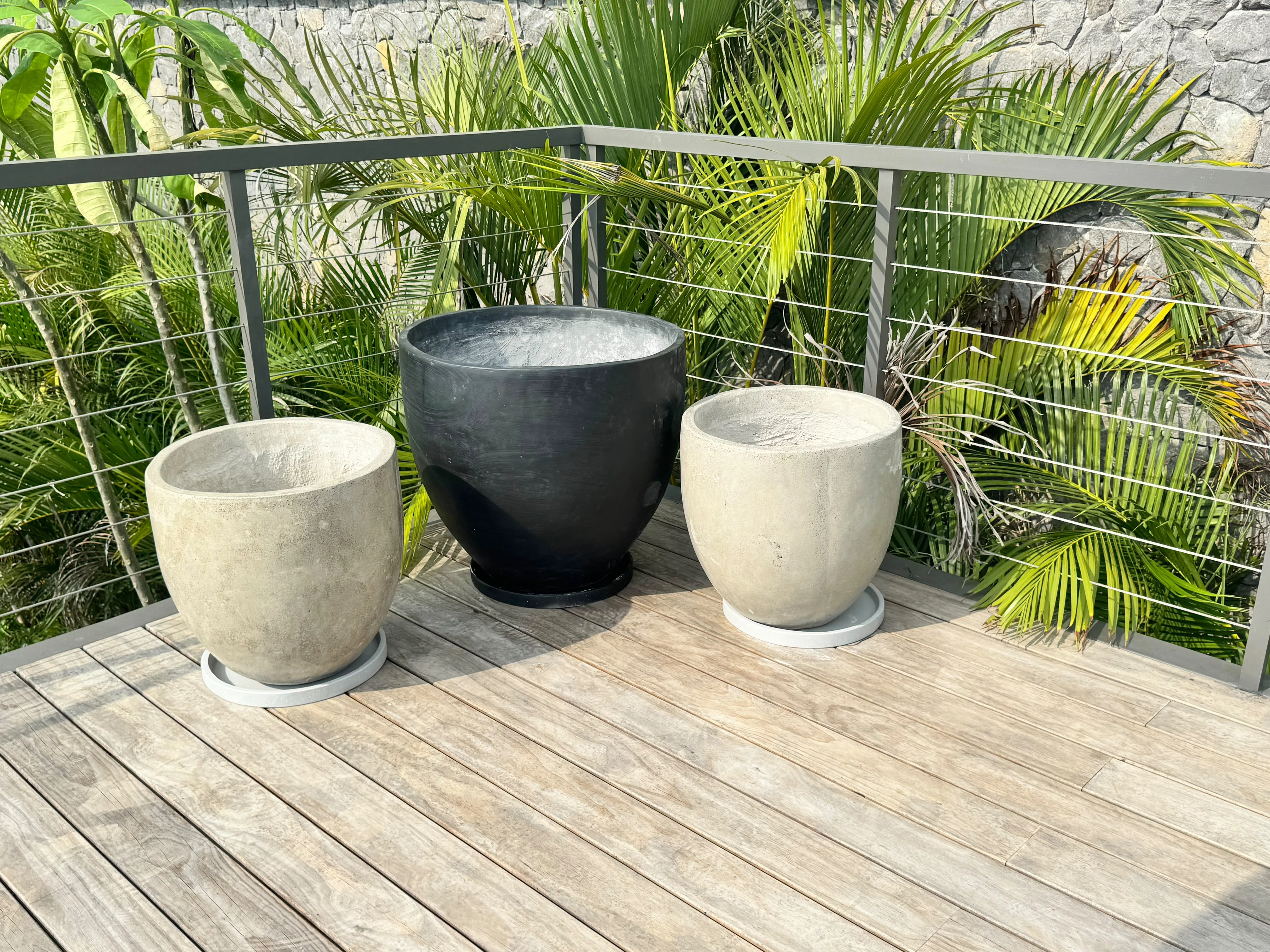 Tres maceteros redondos de concreto, dos de tono claro y uno negro, sobre deck de madera en terraza con vegetación