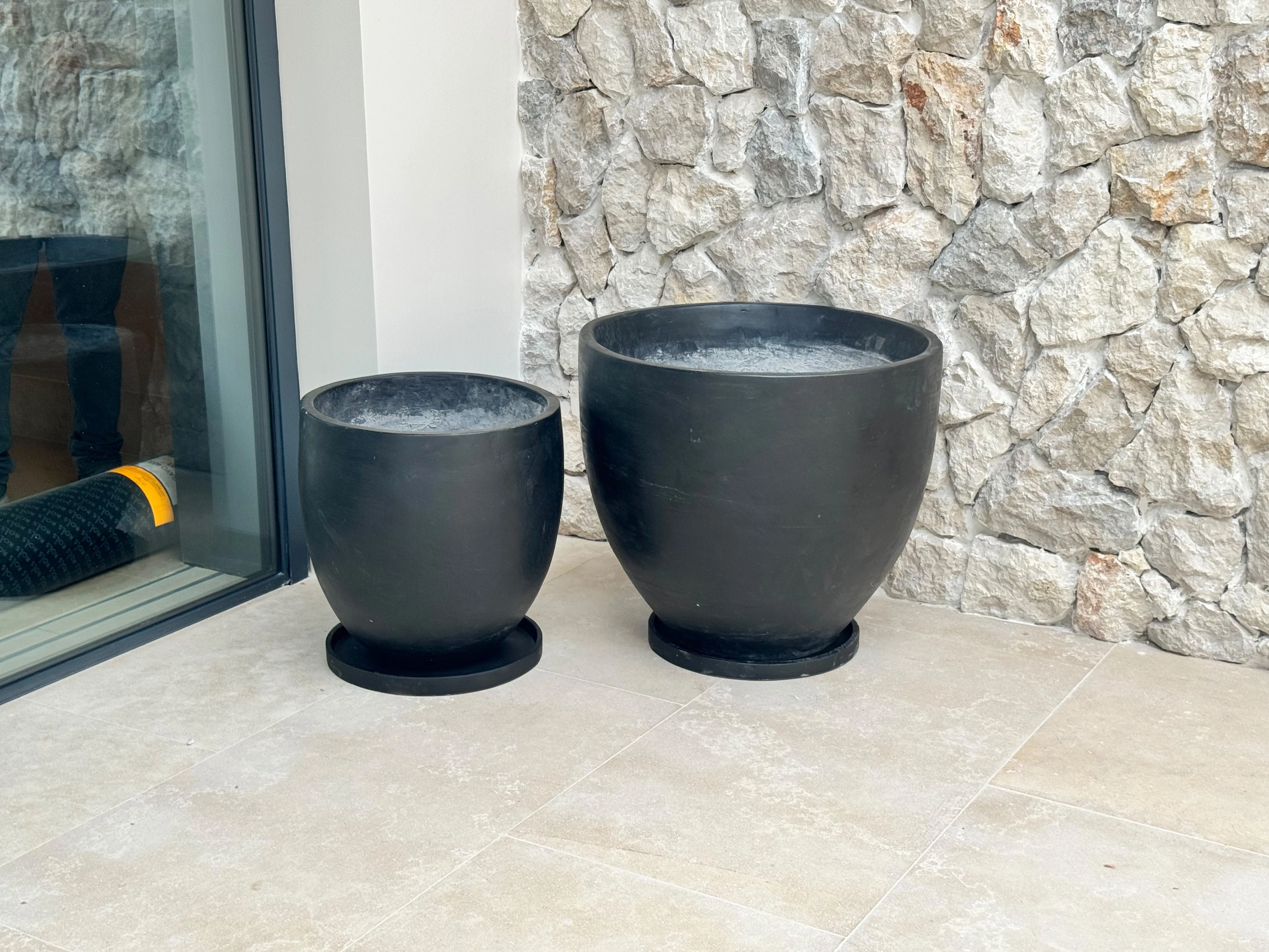 Dos maceteros de concreto negro con forma de copa y platos de base, sobre piso claro junto a pared de piedra