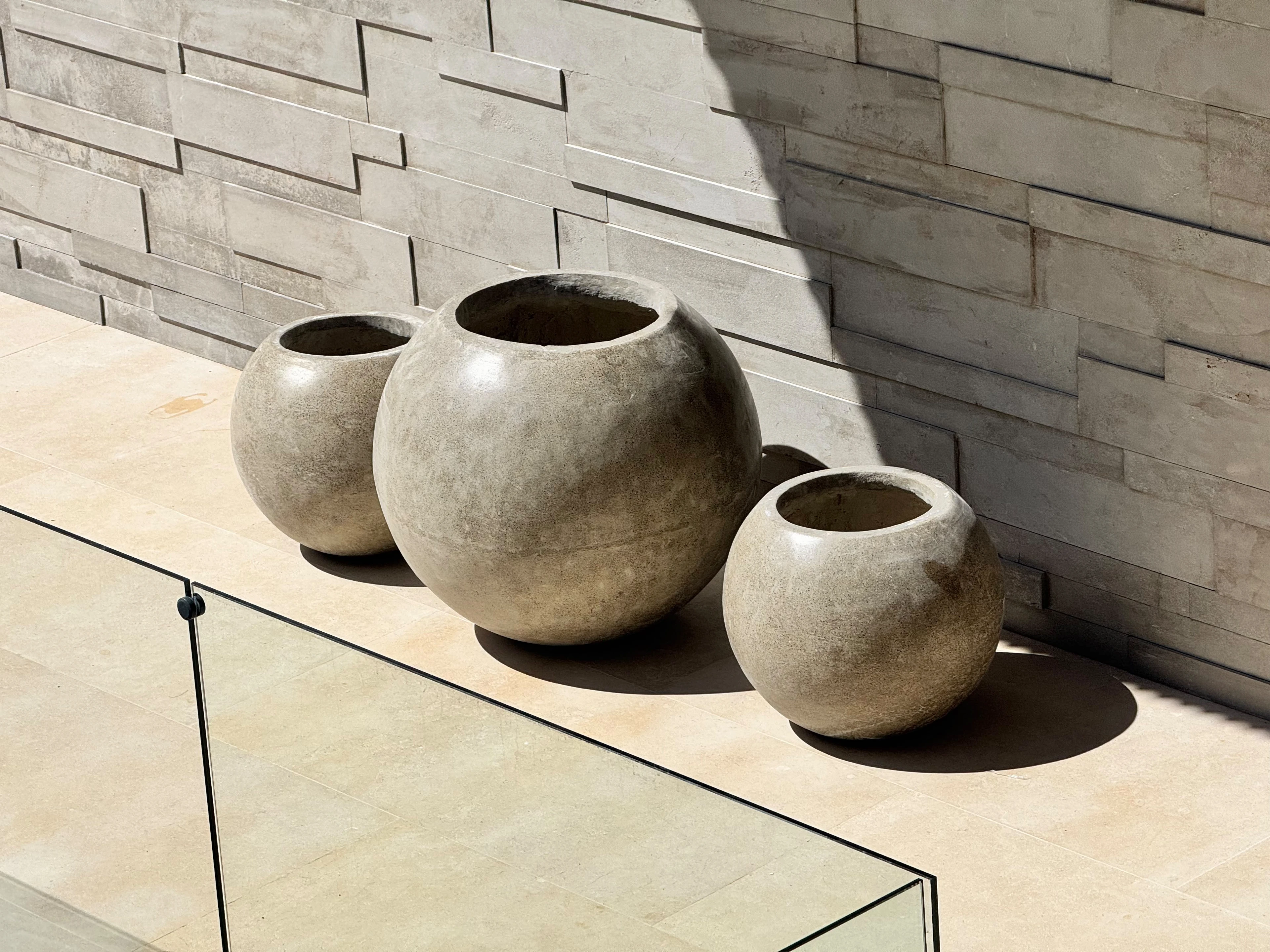 Tres maceteros esféricos de concreto gris de distintos tamaños, sobre terraza con pared de piedra y baranda de vidrio