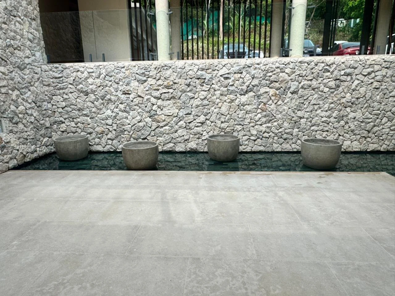 Cuatro maceteros bajos de concreto gris alineados junto a un muro de piedra, sobre un patio con piso de cemento pulido