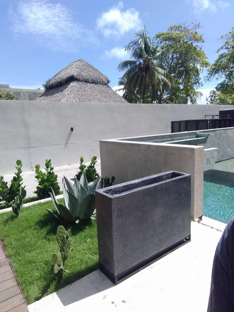 Jardinera alta y angosta de concreto gris oscuro sobre base metálica, en jardín con agave, césped y piscina al fondo
