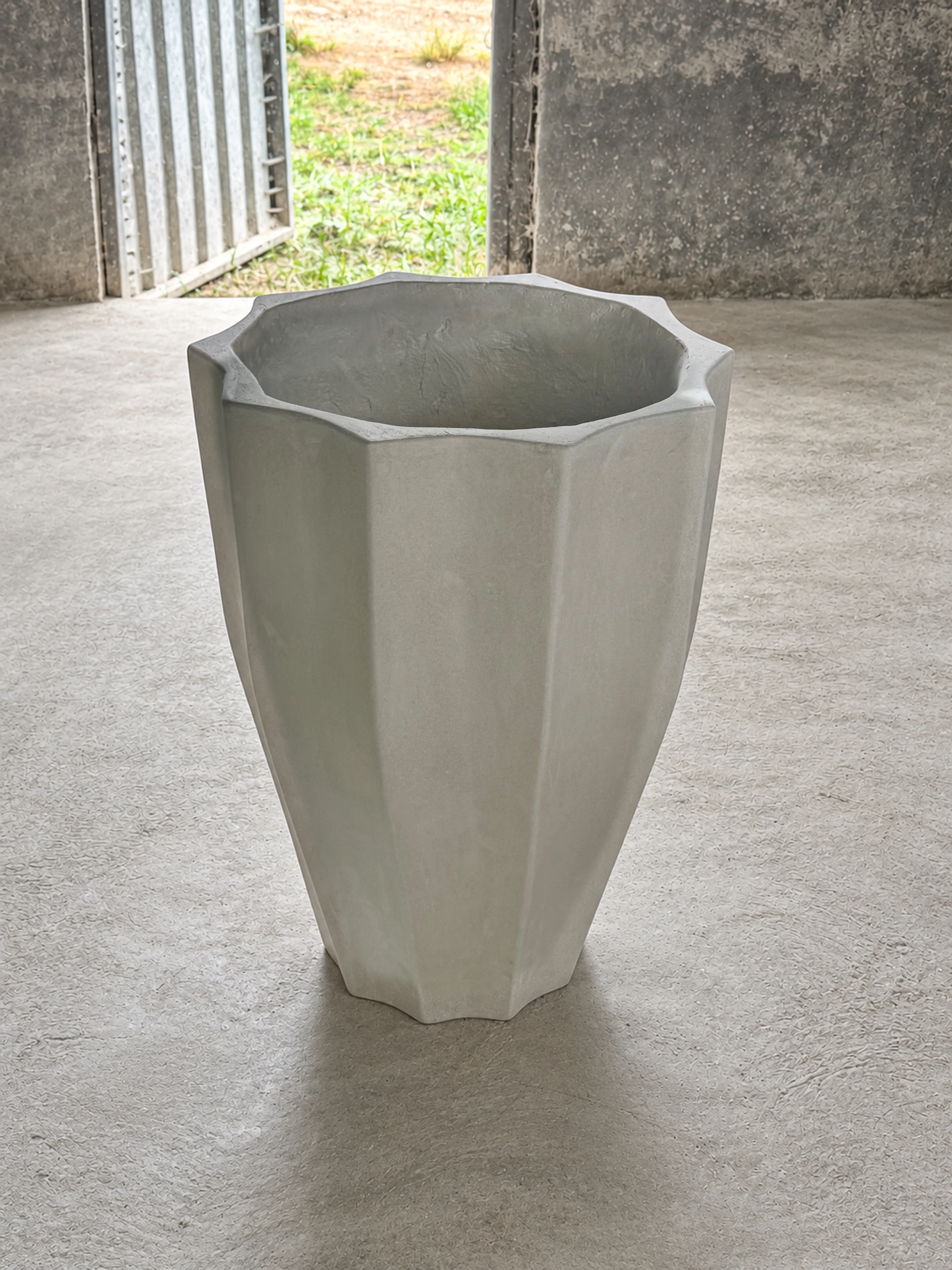 Macetero alto de concreto blanco con paredes facetadas y boca poligonal, sobre piso de cemento en una bodega abierta