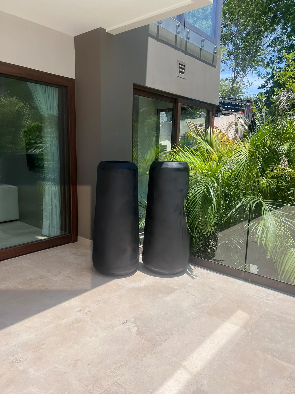Dos maceteros altos y cilíndricos de concreto negro sobre terraza de piedra clara, junto a puertas de vidrio