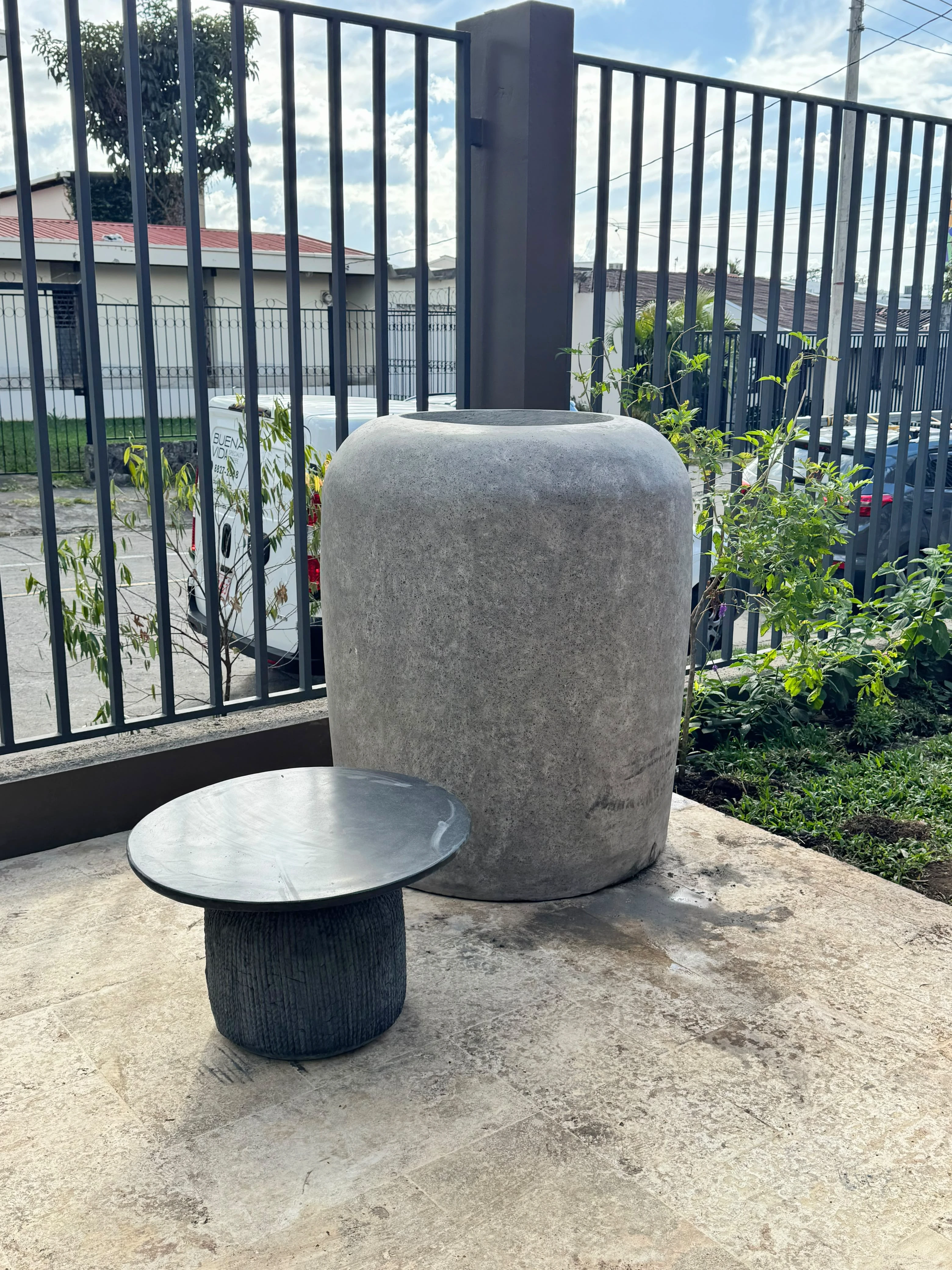 Macetero grande de concreto gris con forma de vasija y una mesa auxiliar redonda oscura, en entrada con reja negra