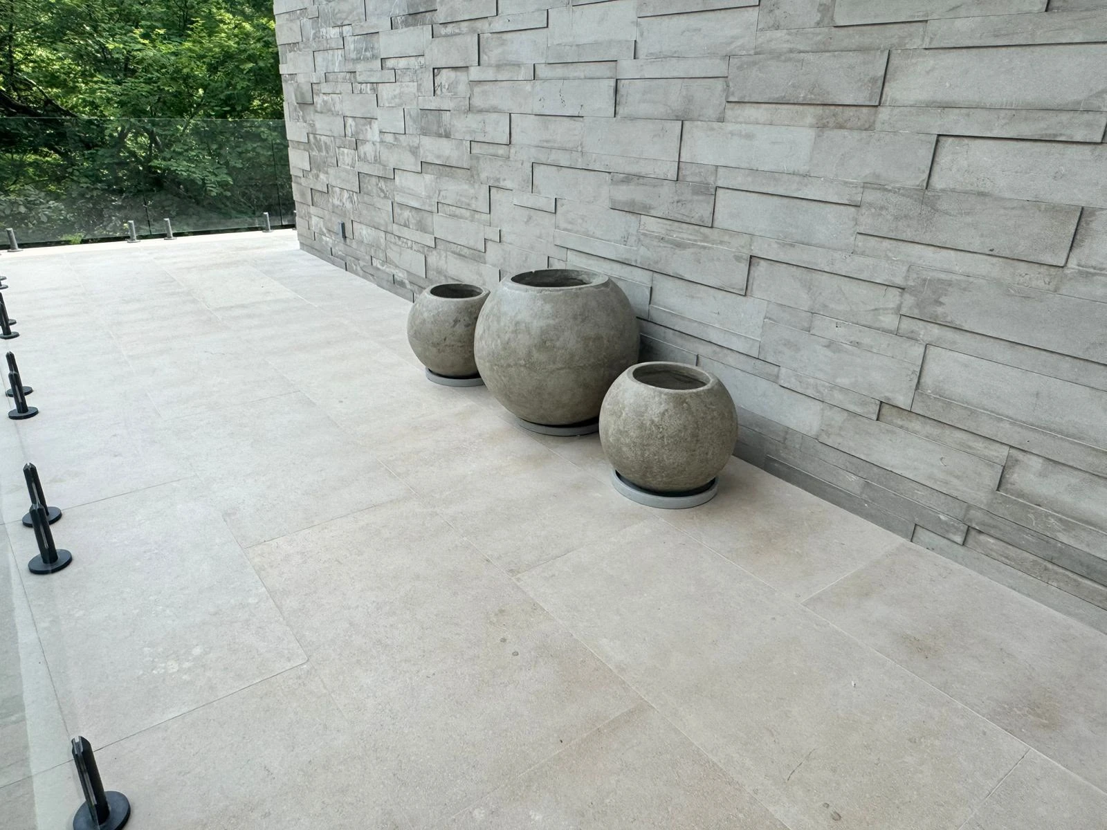 Tres maceteros esféricos de concreto gris con base, sobre terraza de piedra clara junto a un muro de revestimiento gris